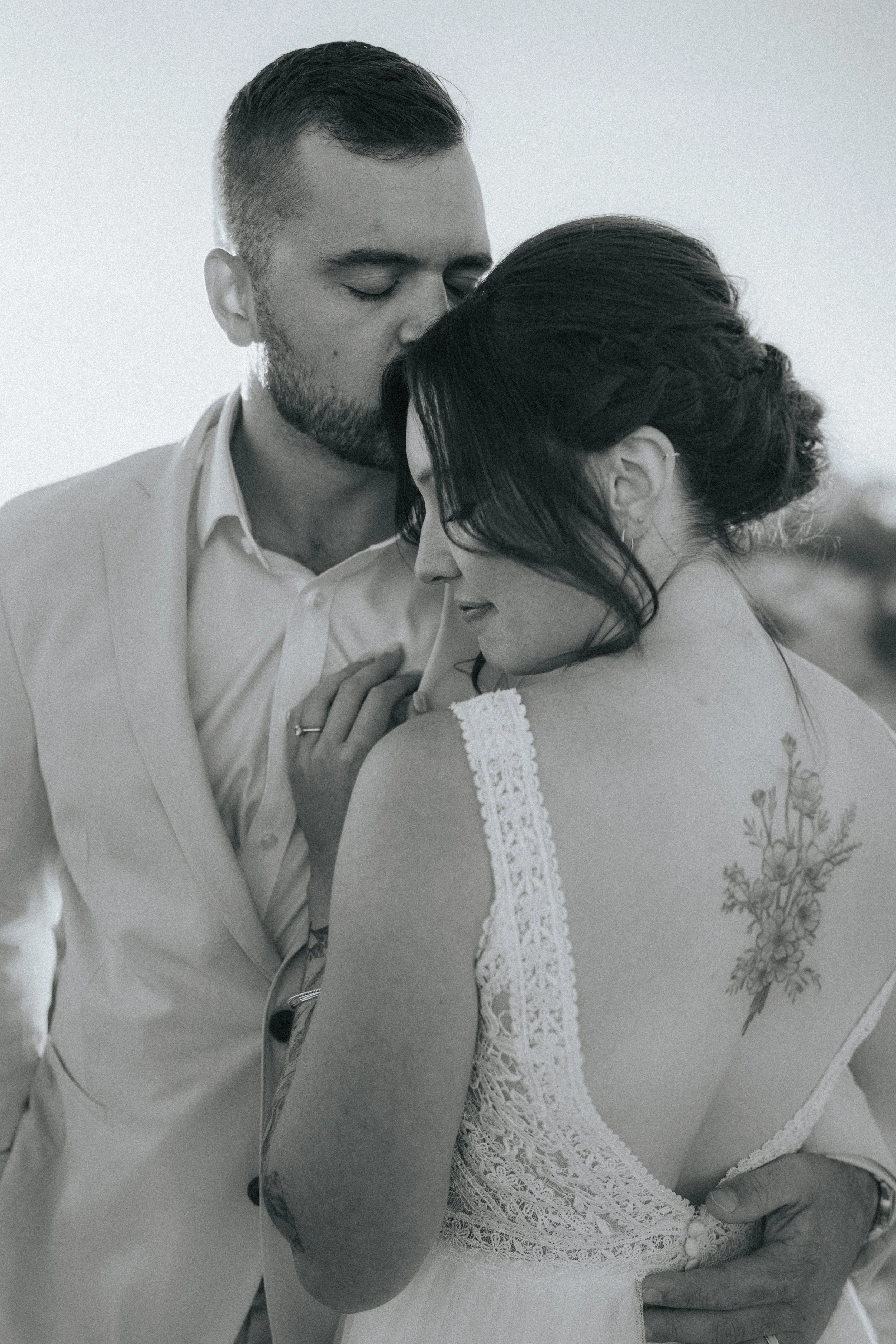 A black and white photograph of a man and woman in an intimate moment. The man has closed eyes, short hair, a beard, and is wearing a light-colored suit. The woman has dark hair styled in an elegant updo, and is wearing a lace dress with floral embro