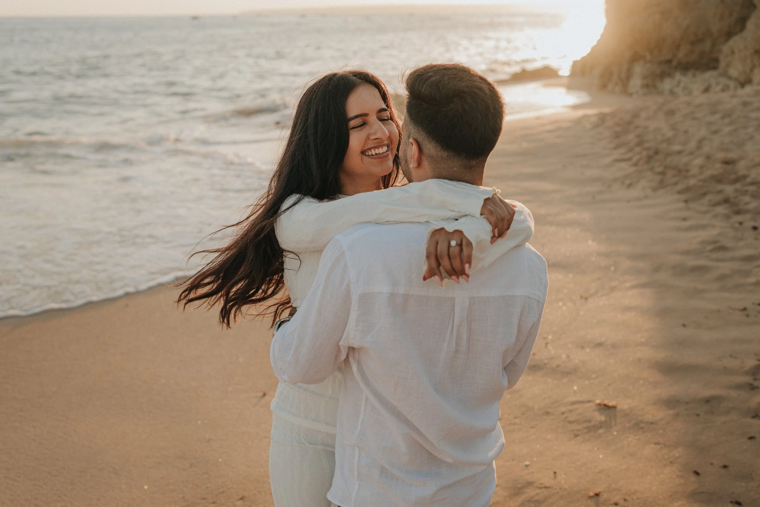 A couple embracing on the beach at sunset. The woman is smiling and has long dark hair, while the man is facing away from the camera. The ocean and cliffs are visible in the background.