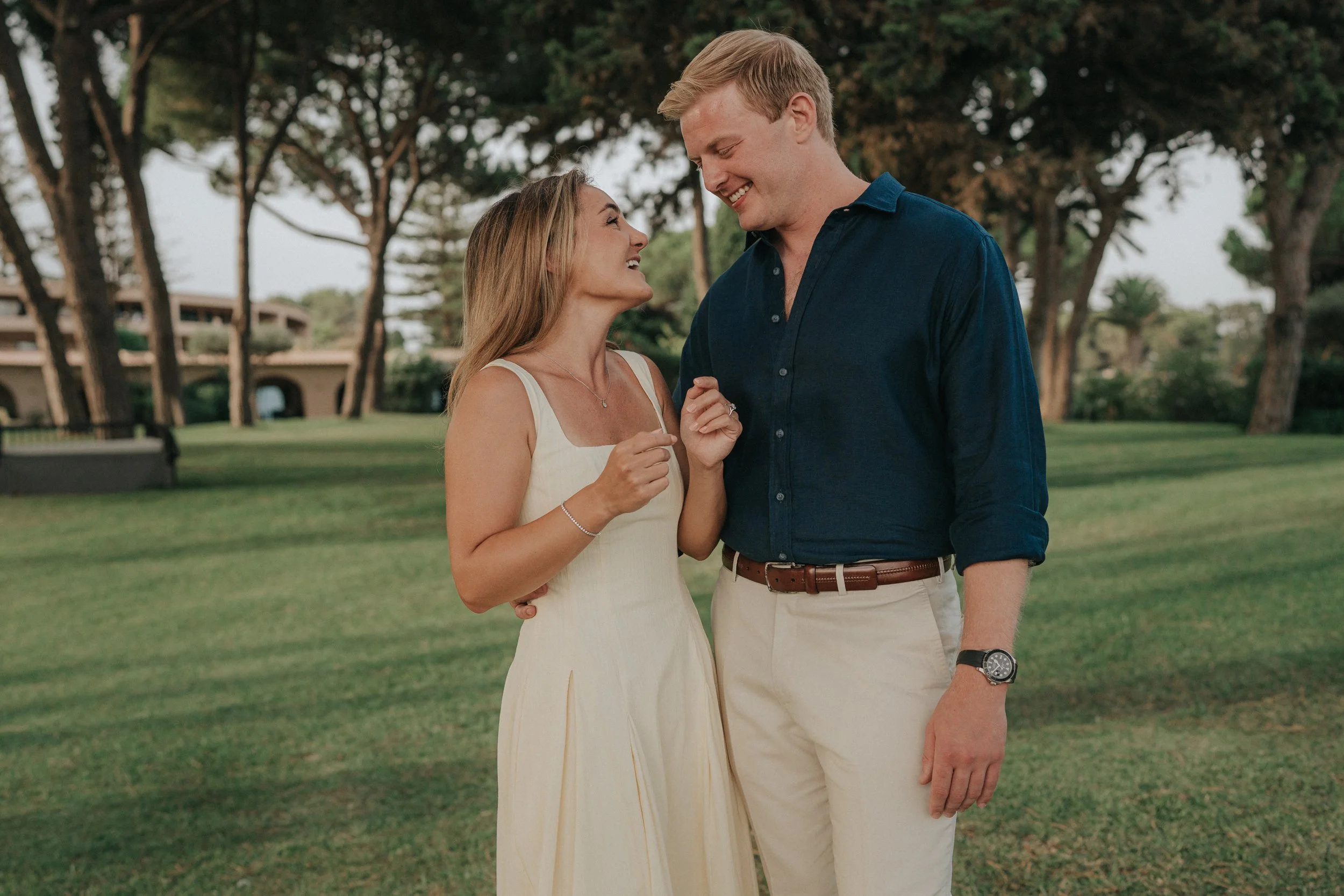 A young woman in a white dress and a young man in a dark blue shirt are smiling and looking at each other affectionately outdoors in a park with trees and green grass.