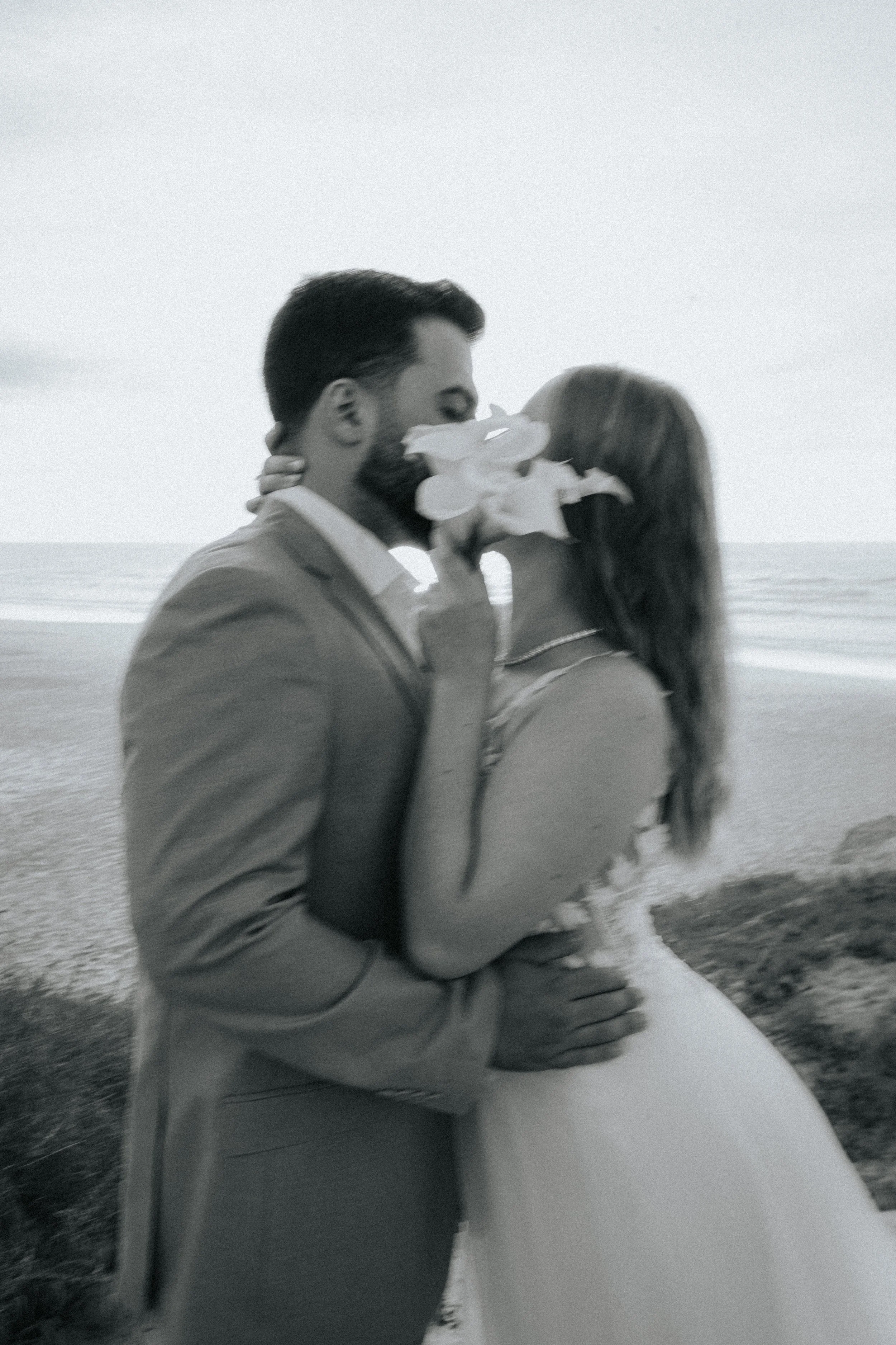 A black and white photo of a couple at the beach, with the man in a suit and the woman in a wedding dress. The woman is sitting on the man's lap, and they are kissing, with the woman's hand on the man's shoulder. The background shows the ocean and th