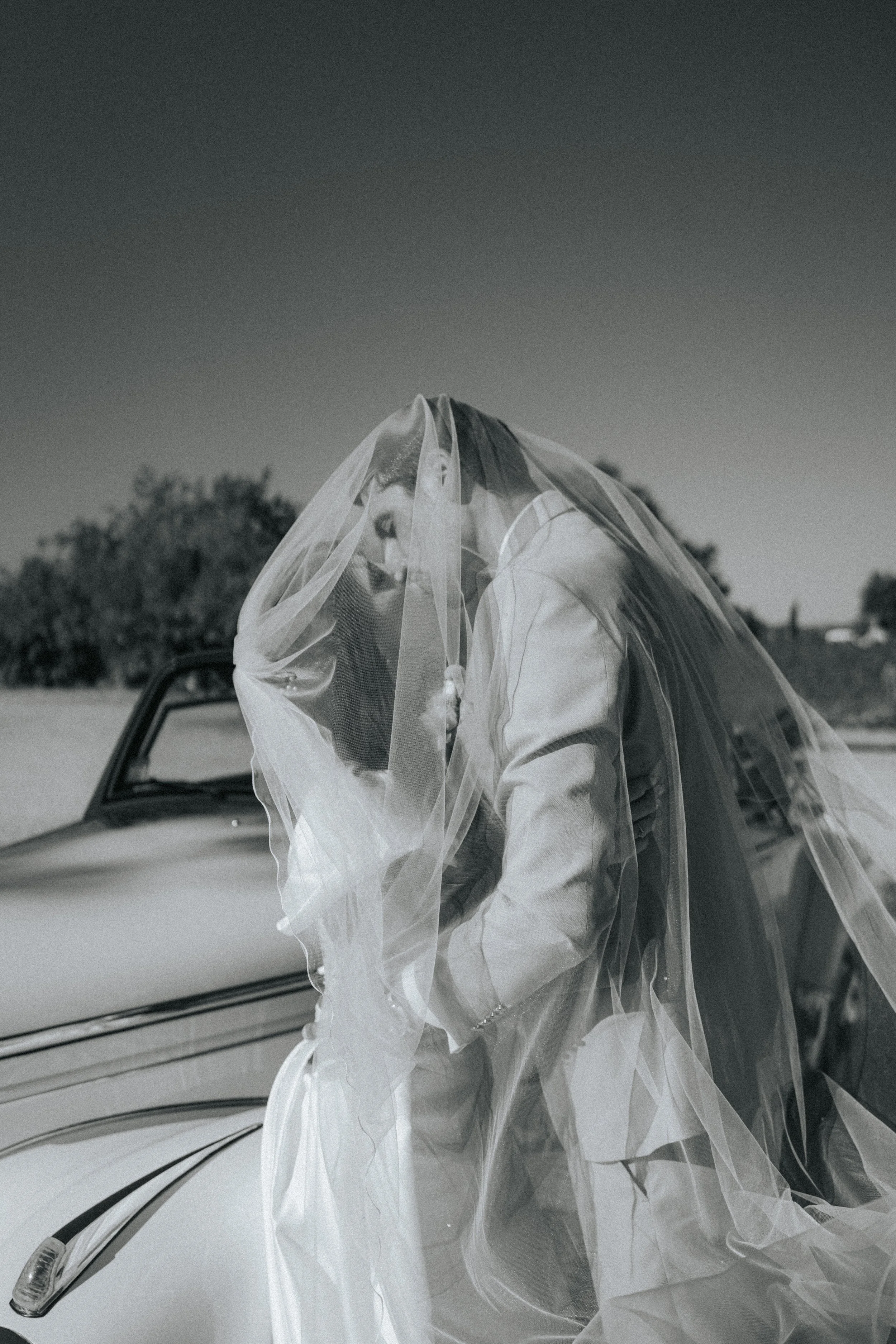 A bride wearing a veil and wedding dress, leaning against a vintage car outdoors.