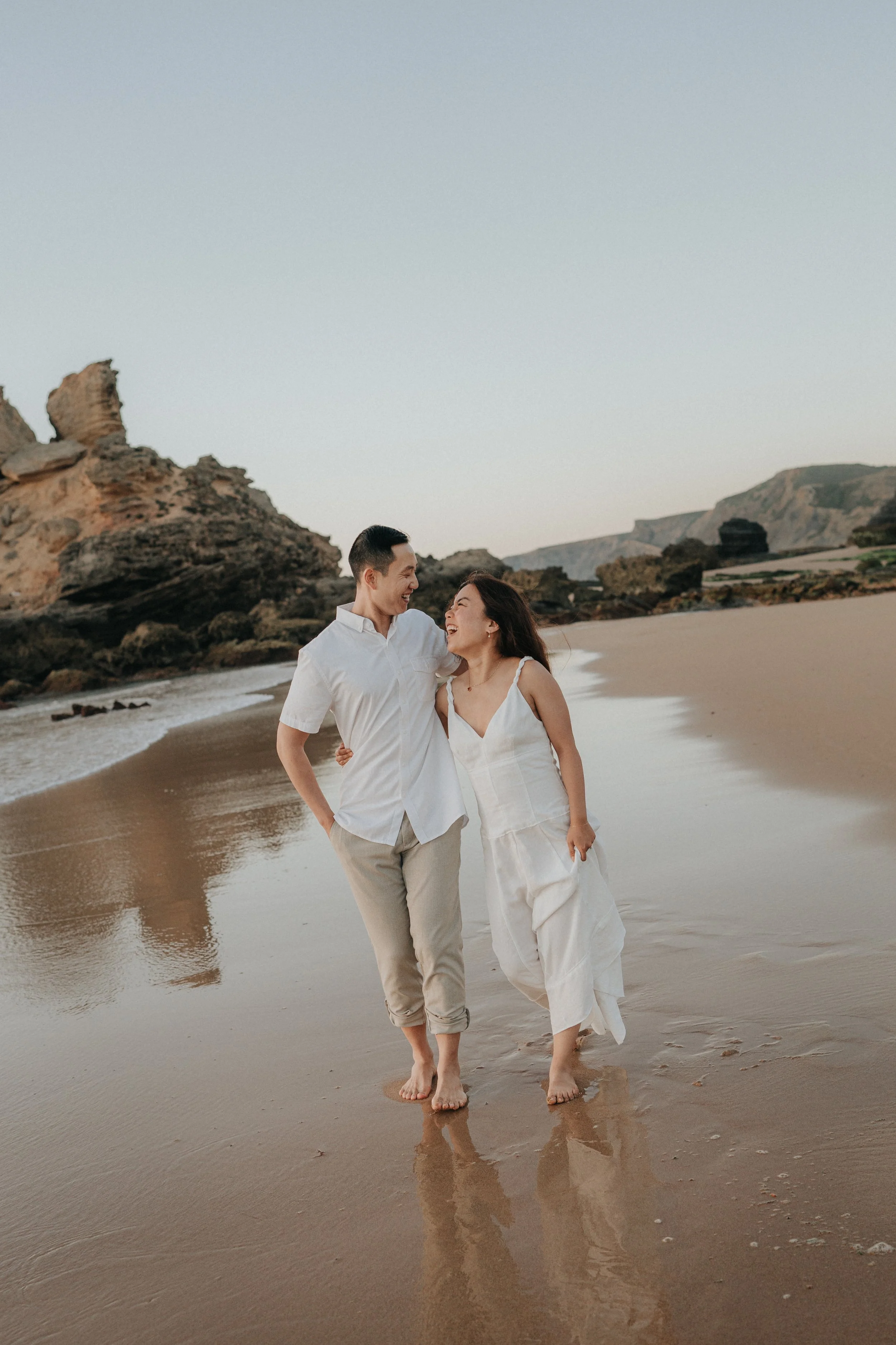 A smiling couple walking on the beach during sunset or sunrise, holding hands and looking at each other, with rocks and cliffs in the background.