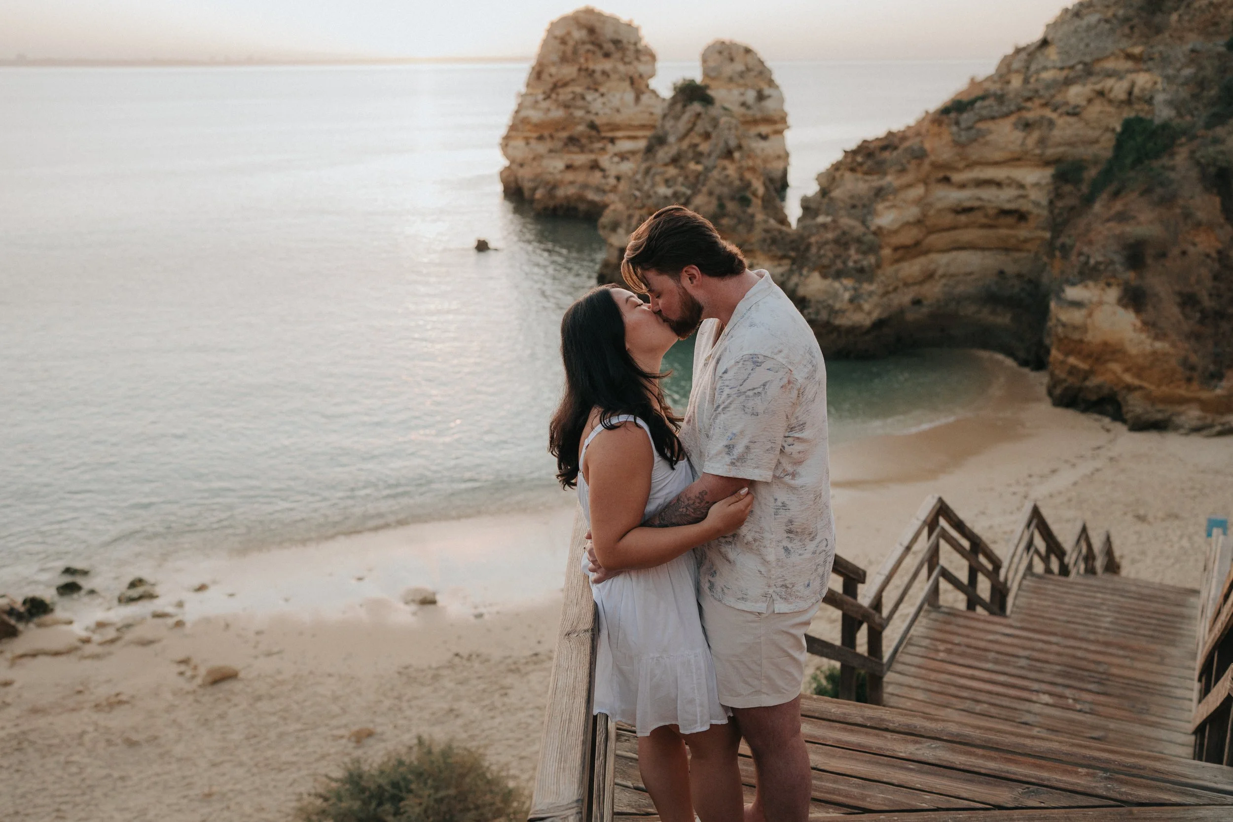 A couple is sharing a kiss on a beach near large rock formations at sunset.