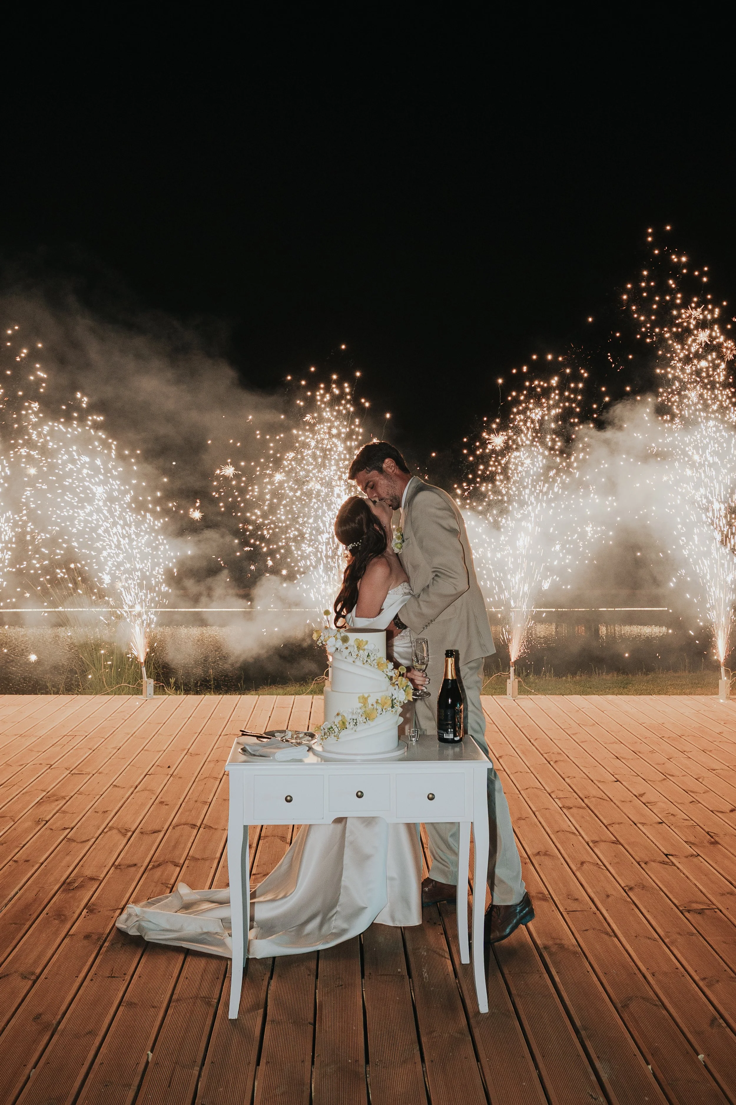 A newlywed couple sharing a kiss during a fireworks display at night, with a wedding cake and champagne on a table in front of them on a wooden deck.