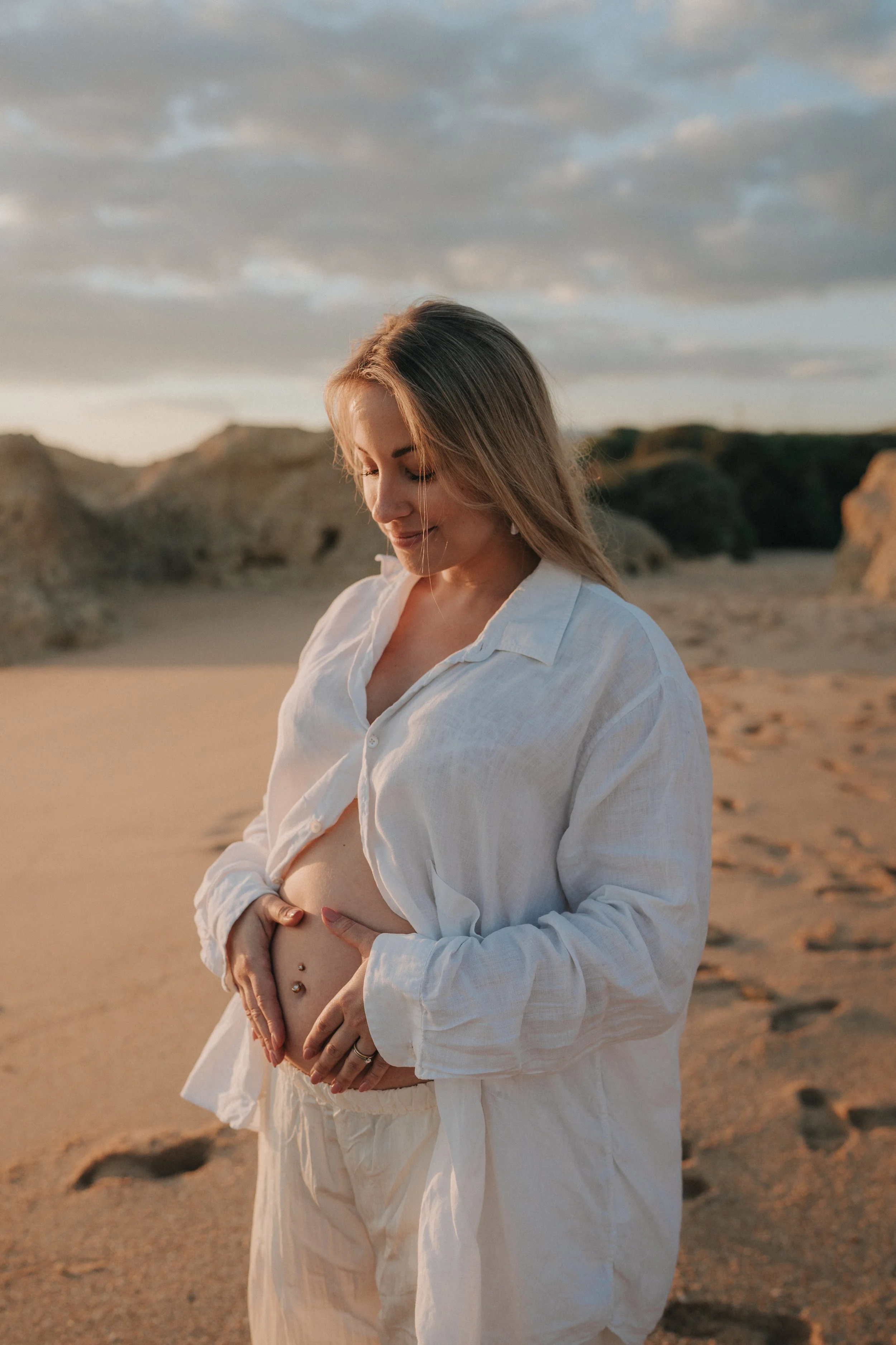 A pregnant woman with a belly piercing stands on a sandy beach during sunset, wearing a white shirt and pants, with her hands gently cradling her belly and a serene smile on her face.