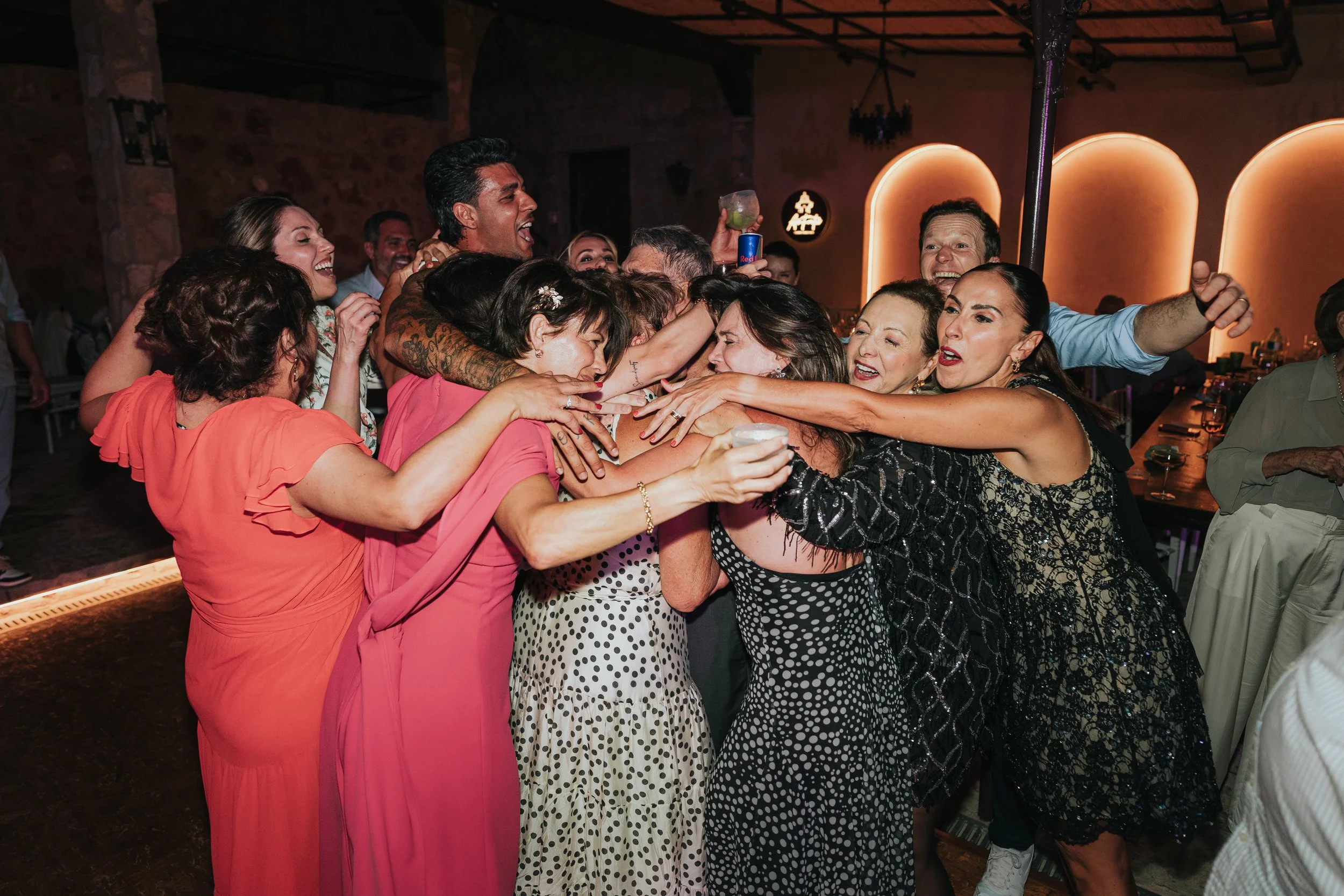 A group of people hugging and celebrating at a lively party, some holding drinks and smiling.