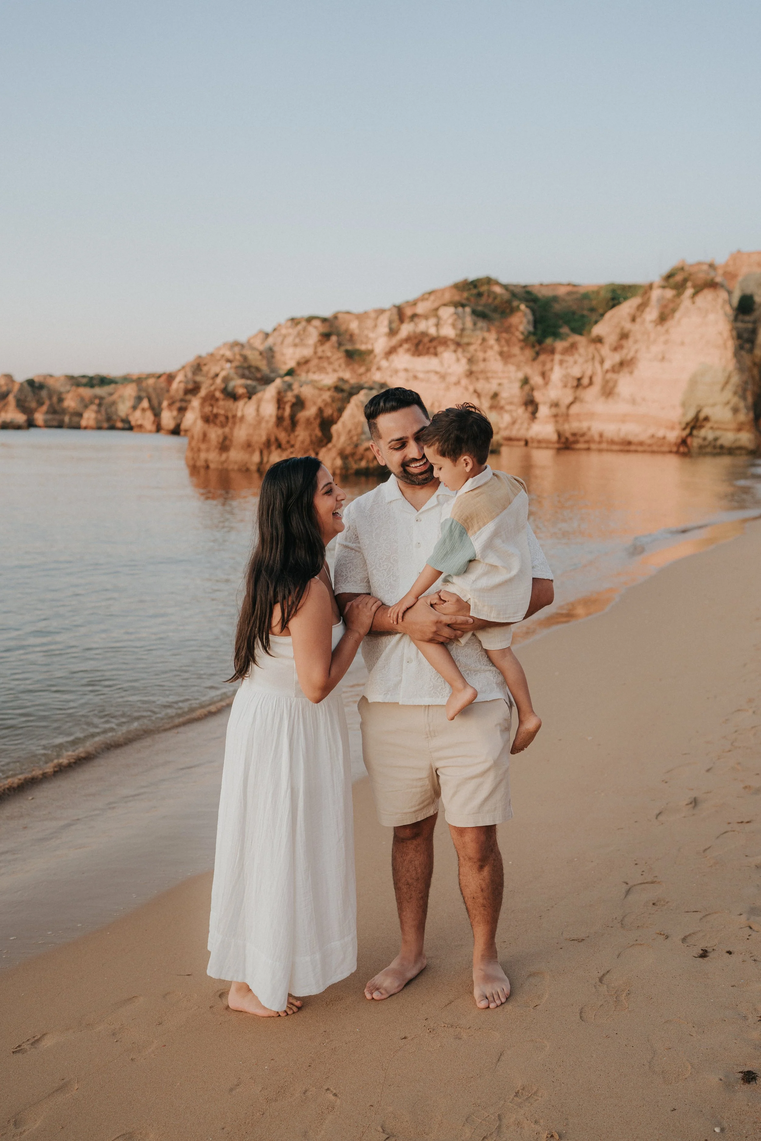 A family of three standing on the beach during sunset, with rocky cliffs in the background. The woman has long dark hair and is wearing a white dress, the man is smiling and holding a young boy, who is wearing a light-colored shirt and shorts. They a