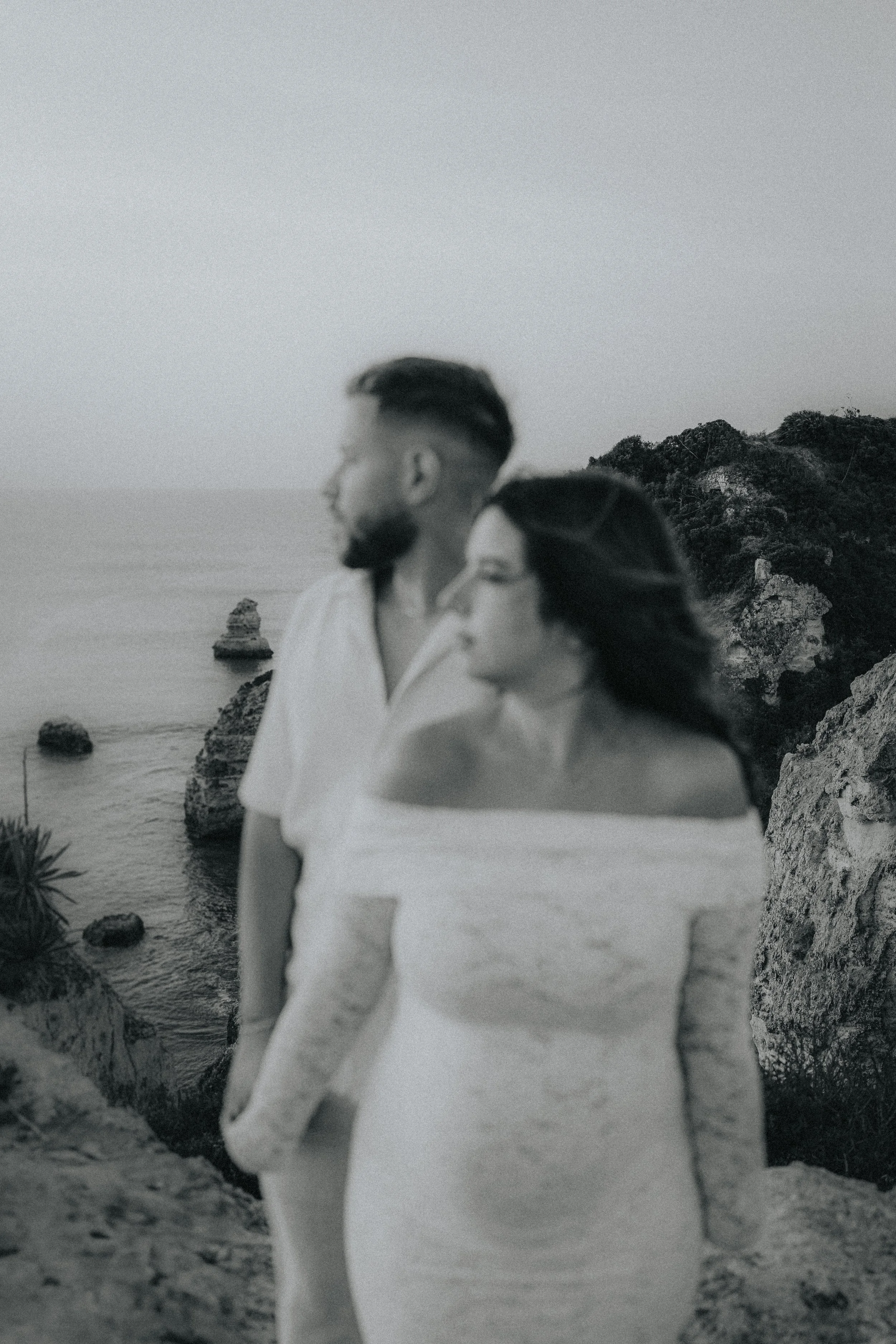 A black and white photo of a man and woman standing on a rocky coastline, overlooking the ocean with cliffs in the background. The man has a beard and styled hair, and is looking away, while the woman has long hair and is looking down.