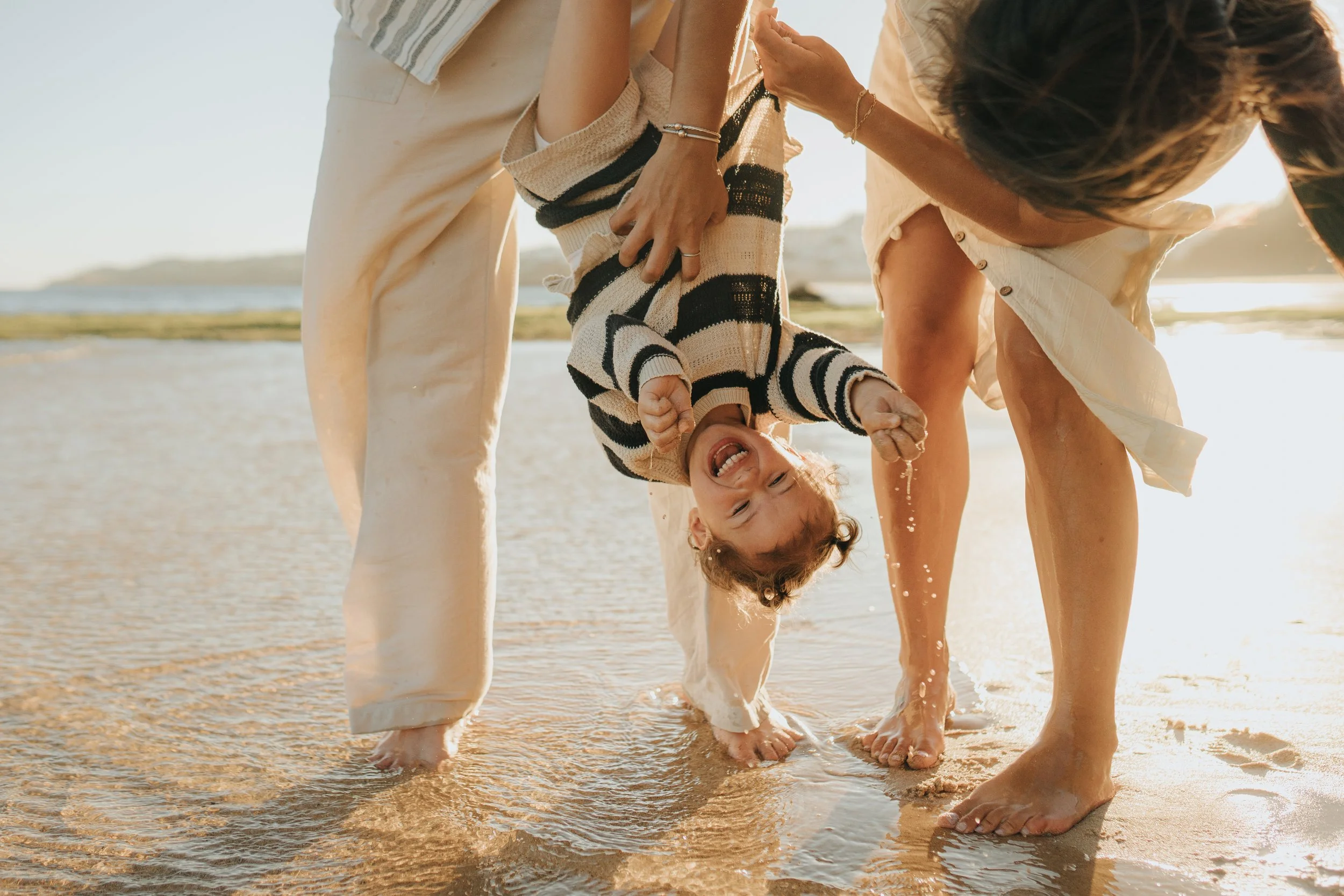 Family walking in shallow water at the beach, holding a child upside down, with the sun setting or rising.