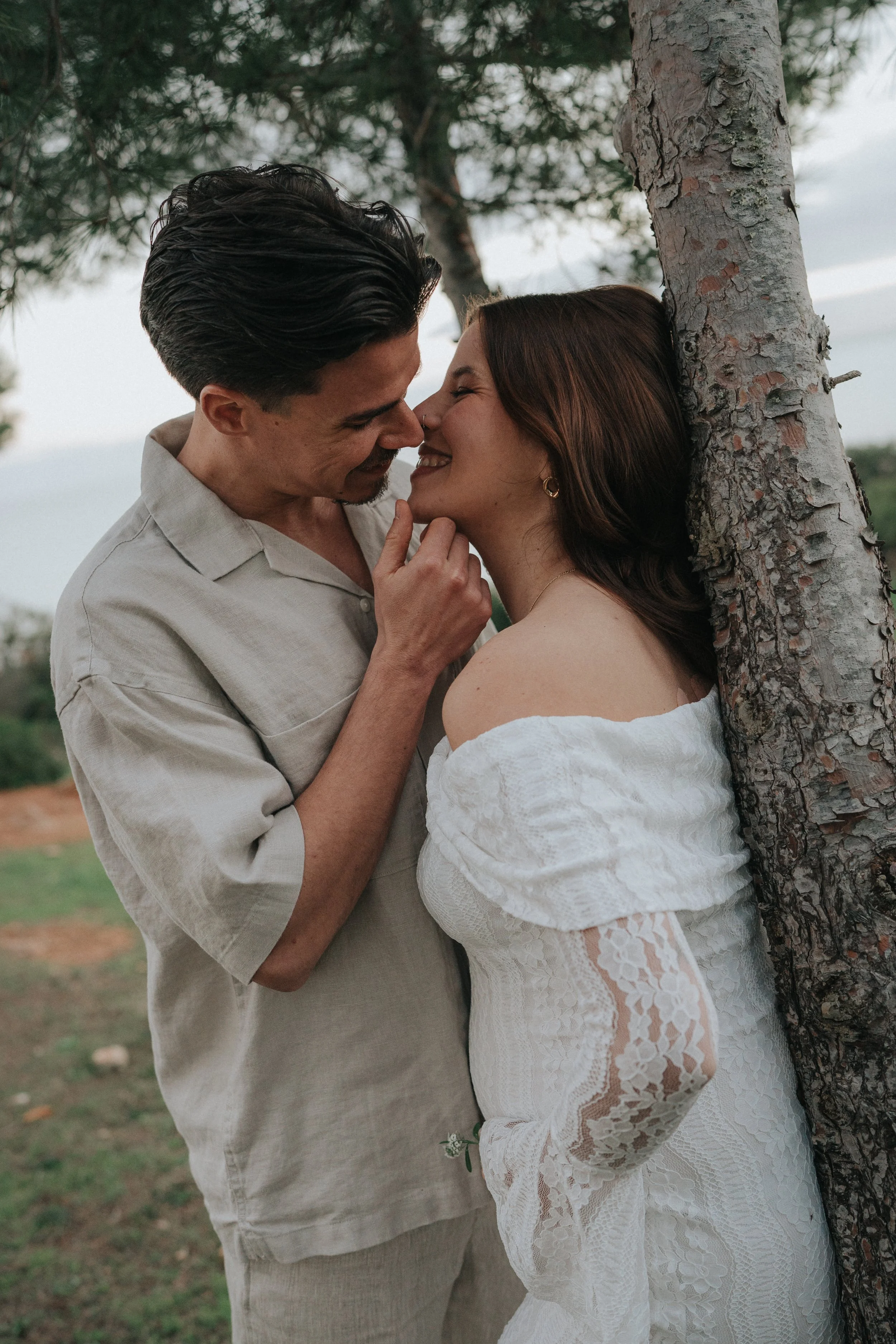 A man and woman are close to each other, touching noses and smiling, with the woman leaning against a tree outdoors in a natural setting.