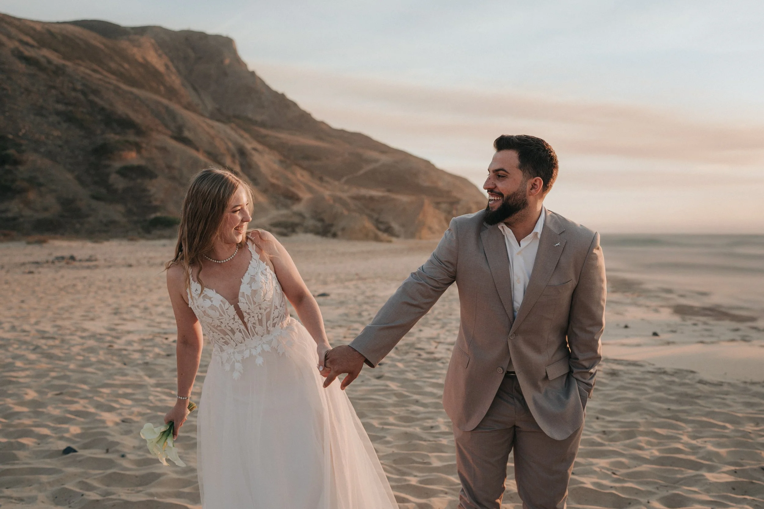 A bride and groom holding hands and smiling on the beach at sunset, with cliffs in the background.