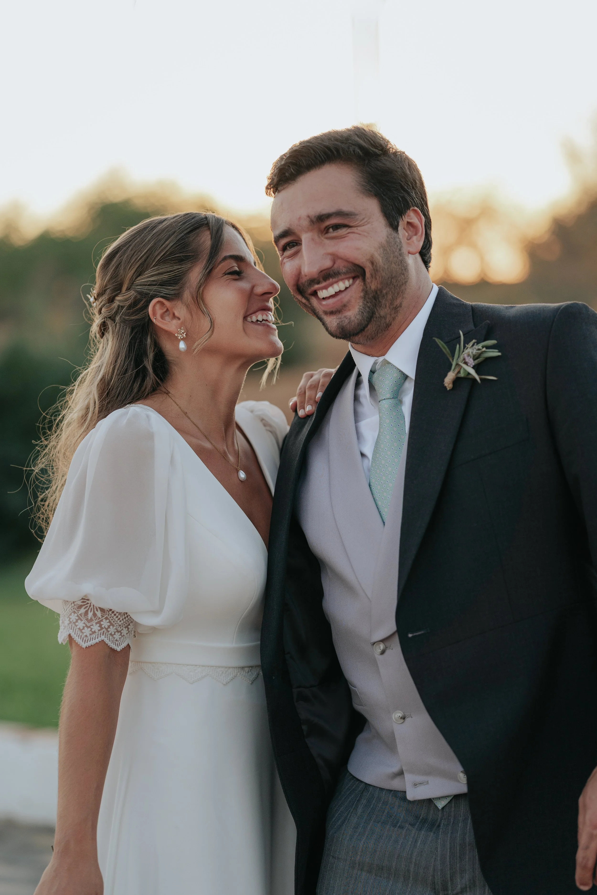 A smiling couple dressed in wedding attire outdoors at sunset, with the woman in a white dress and the man in a tuxedo, embracing each other and looking happy.