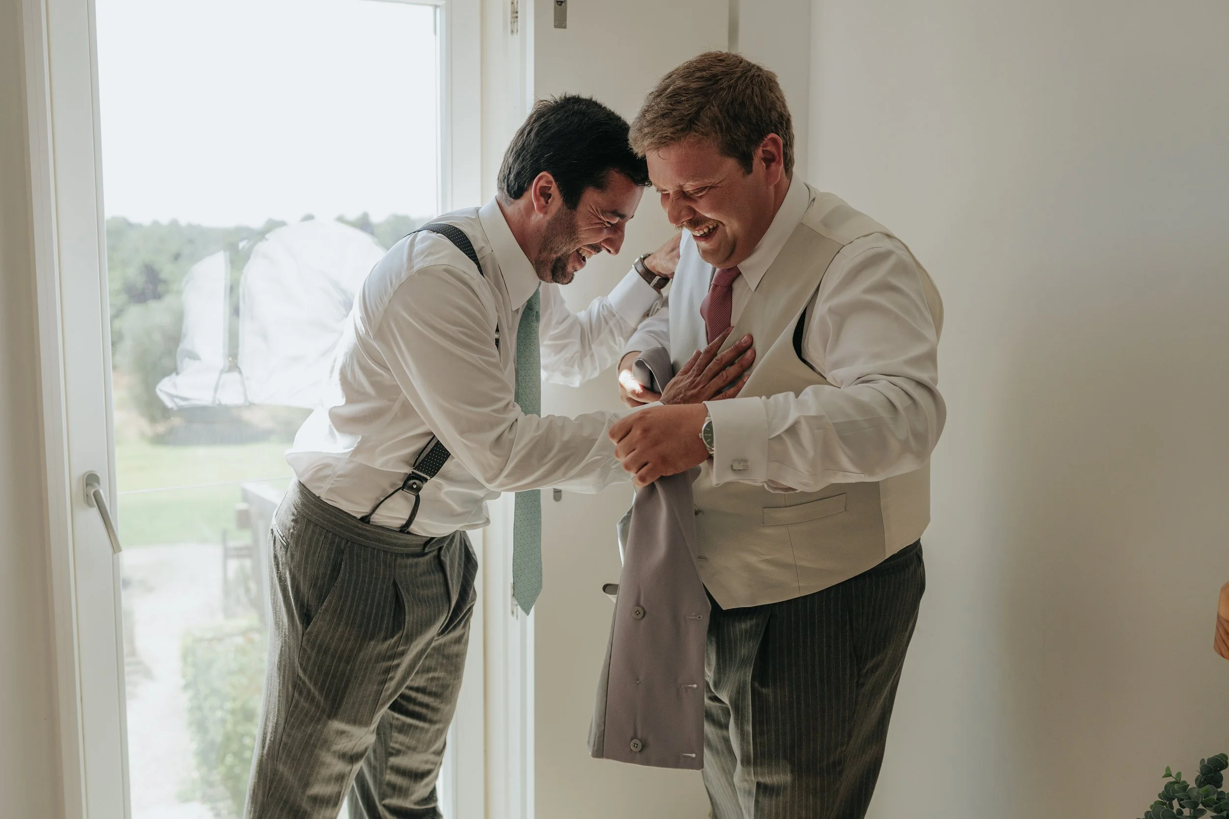 Two men in formal attire, smiling and laughing as one helps the other with his tie inside a bright room near a window.