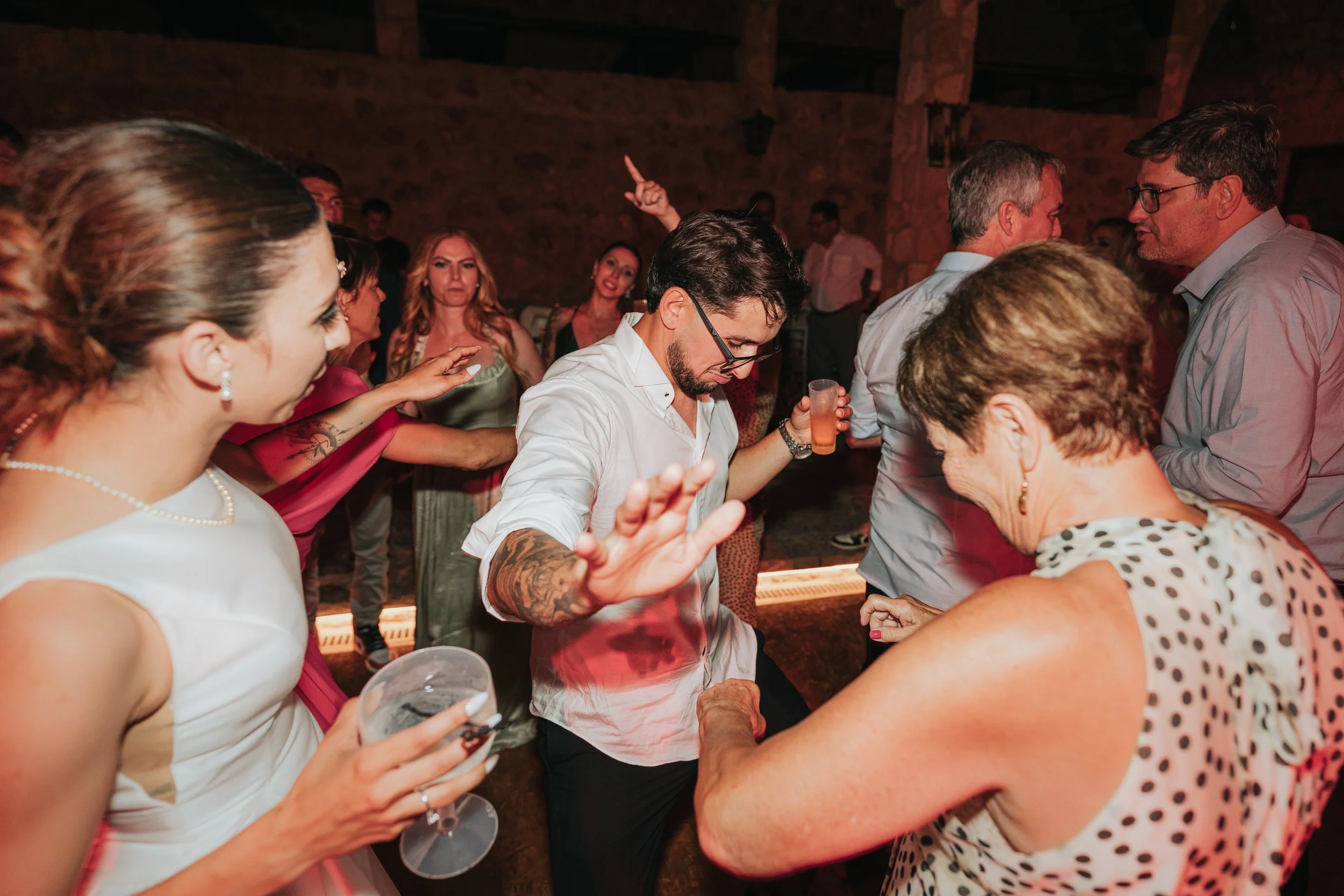 People dancing and socializing at a party with a rustic brick wall background, some holding drinks and engaging in conversations.