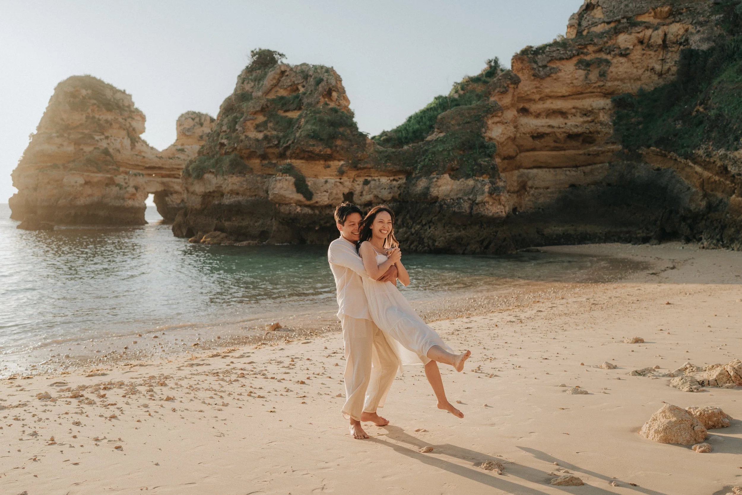 A couple standing on a sandy beach near rock formations, with the woman being lifted by the man and both smiling.