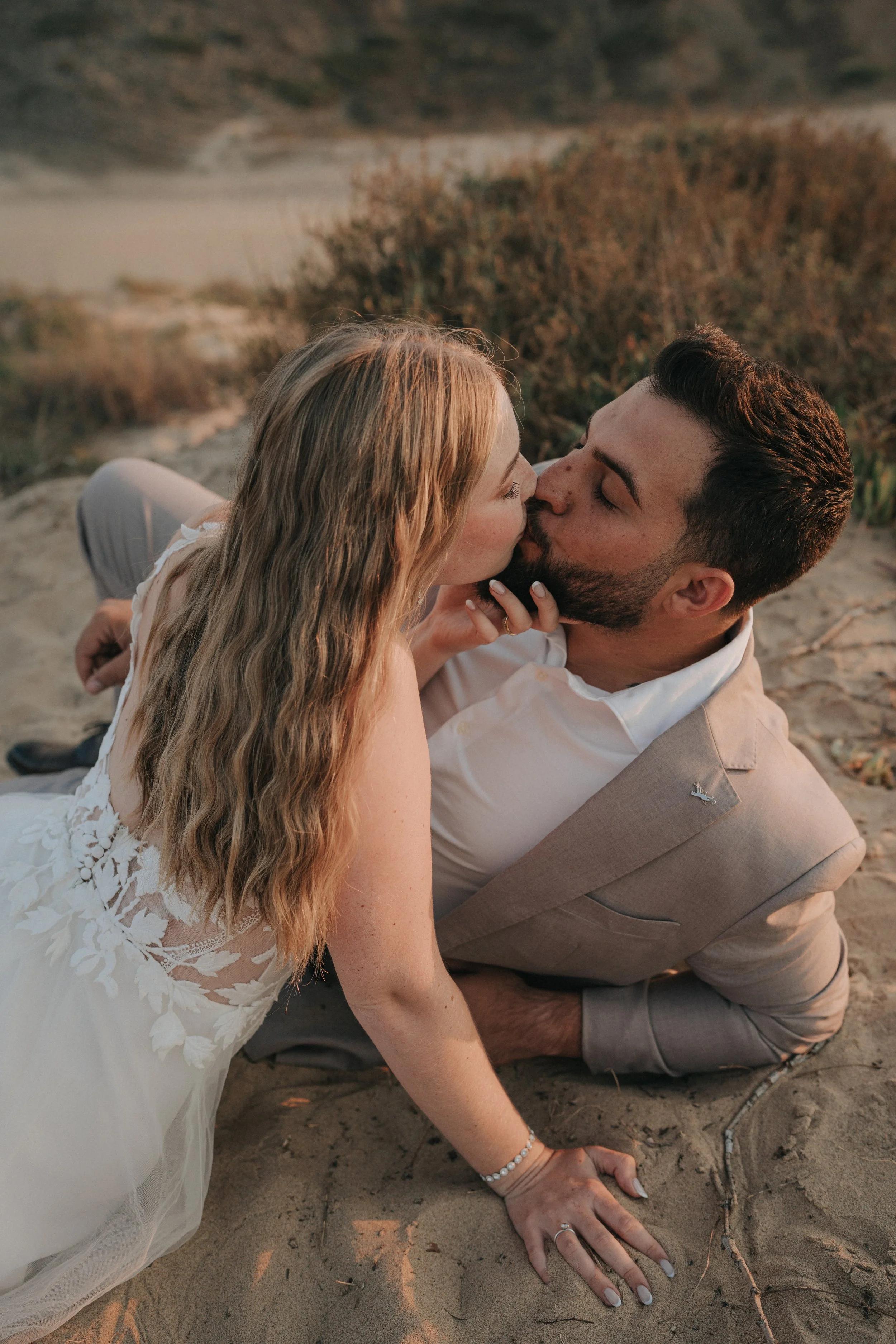 A couple sharing a kiss on the beach, with the woman's hand and jewelry visible on the sandy ground.