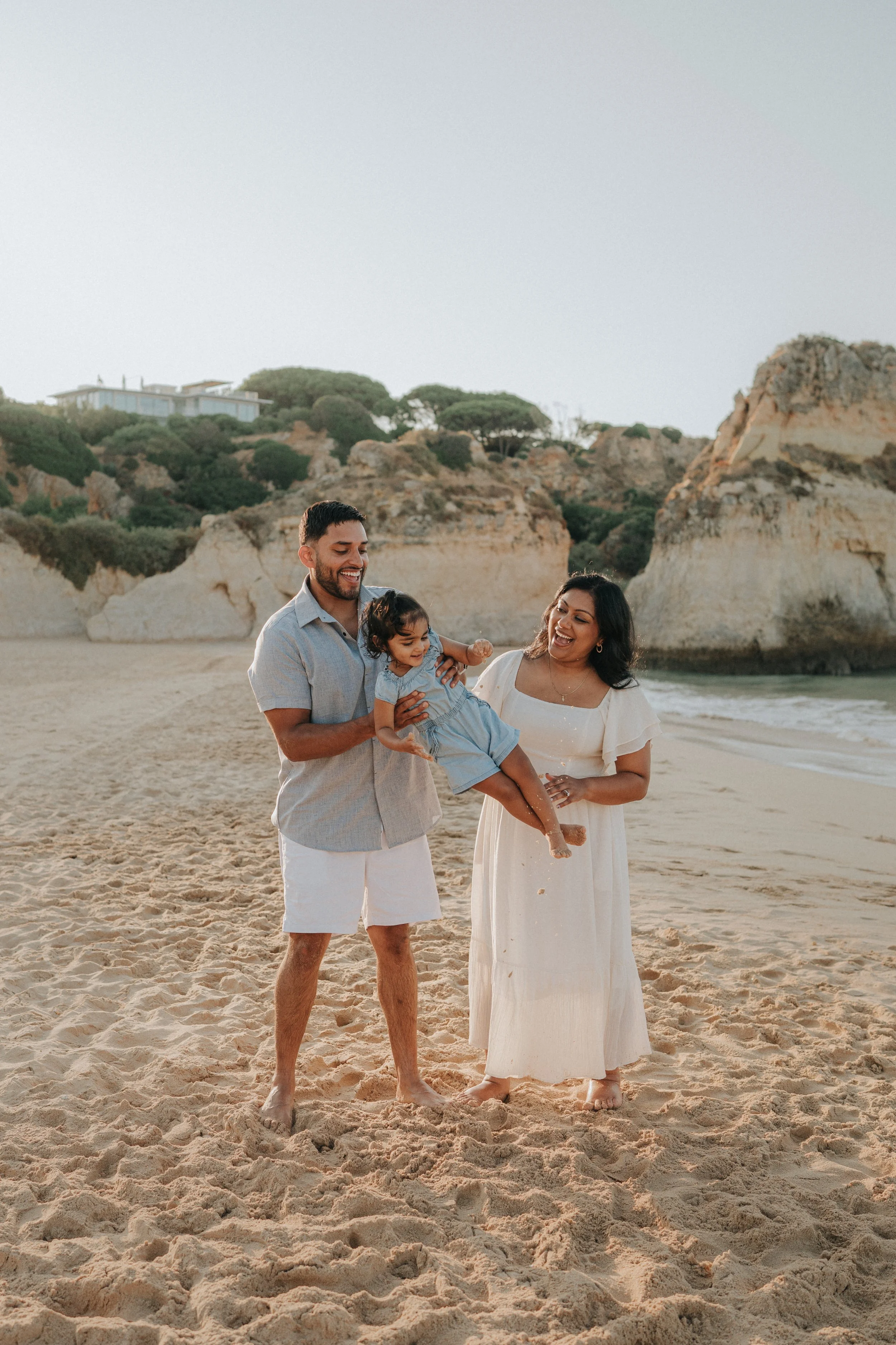 A happy family of three enjoying time on a sandy beach with rocky cliffs and greenery in the background, during the daytime. The father, mother, and child are smiling and smiling, with the child being held by the father.