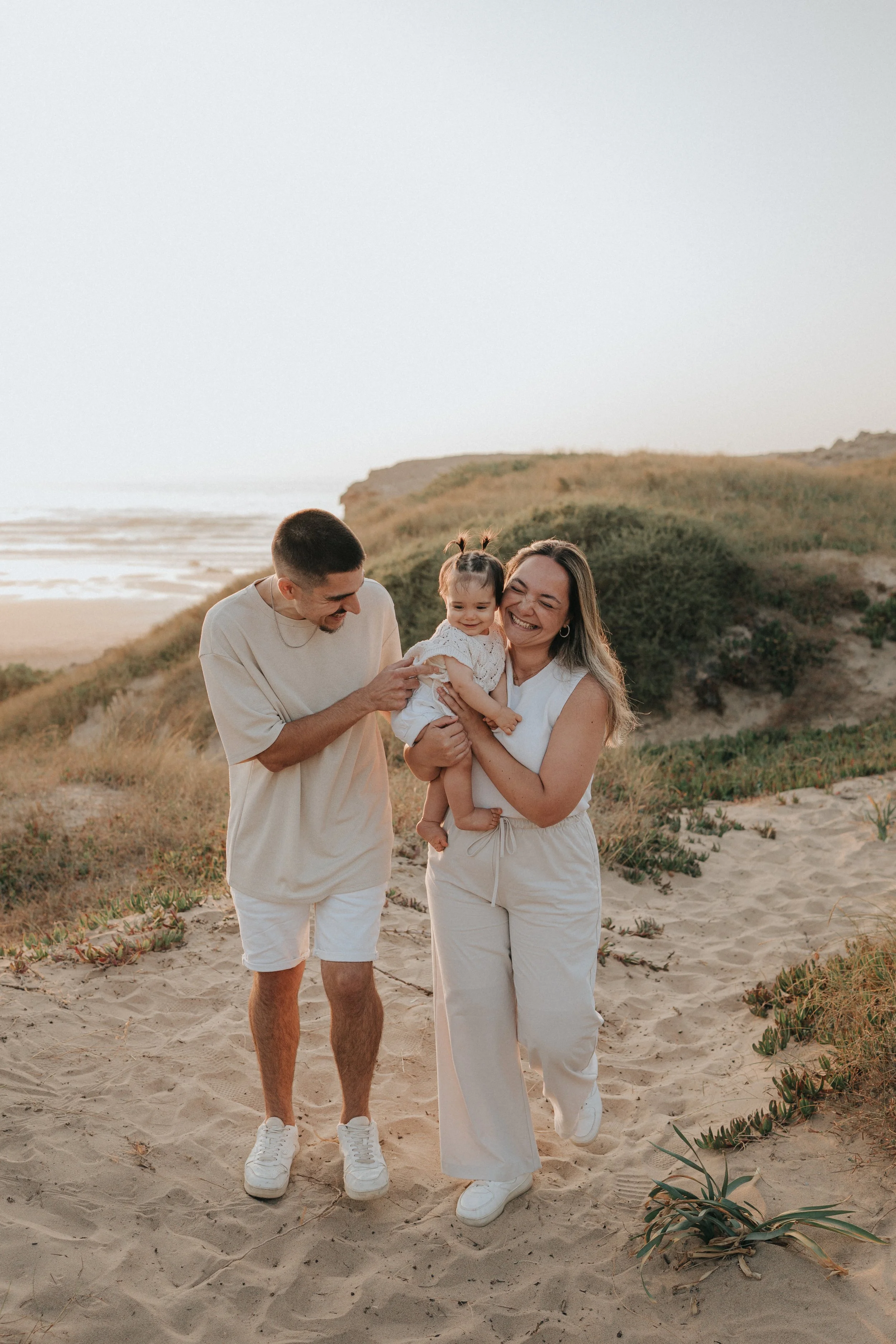 A family of three enjoying a walk on the beach during sunset. The man, woman, and young girl are smiling and holding each other, dressed in light-colored casual clothing. The sandy beach and coastal vegetation are visible in the background.