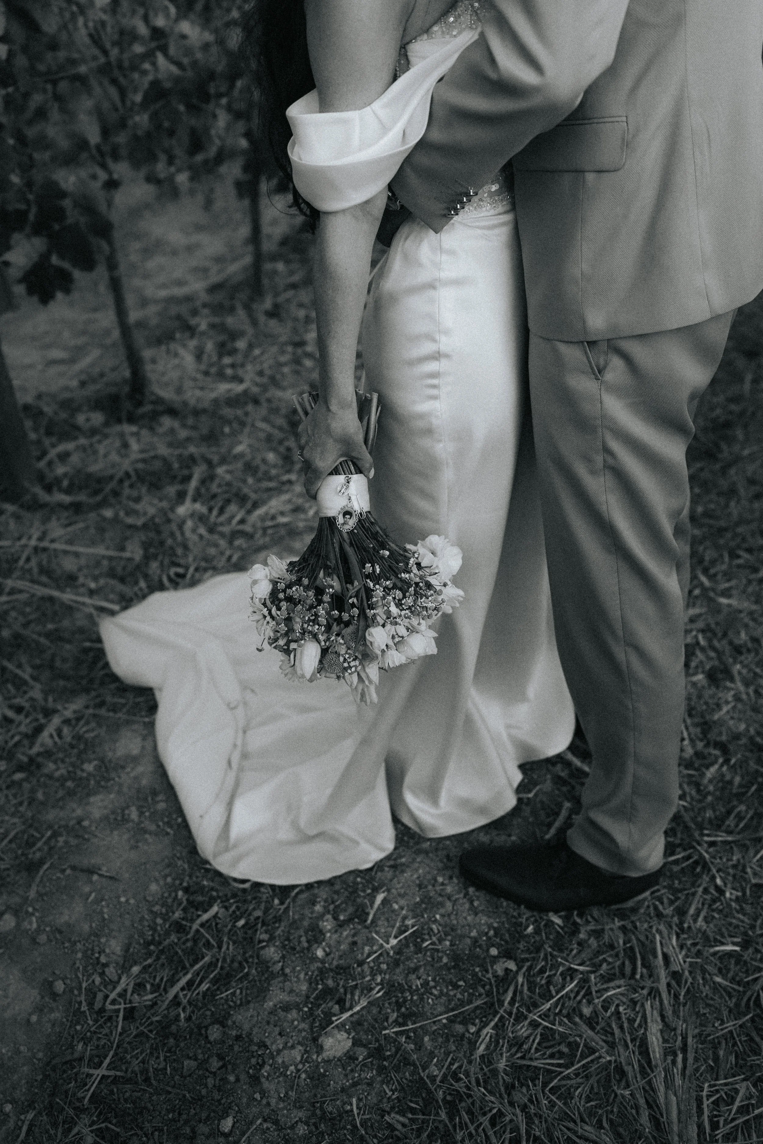 A close-up black-and-white photo of two people, one wearing a suit and the other in a dress, holding a bouquet of flowers. The person in the dress is bent over with their hand reaching down, and the scene appears to be outdoors on a dirt ground with 