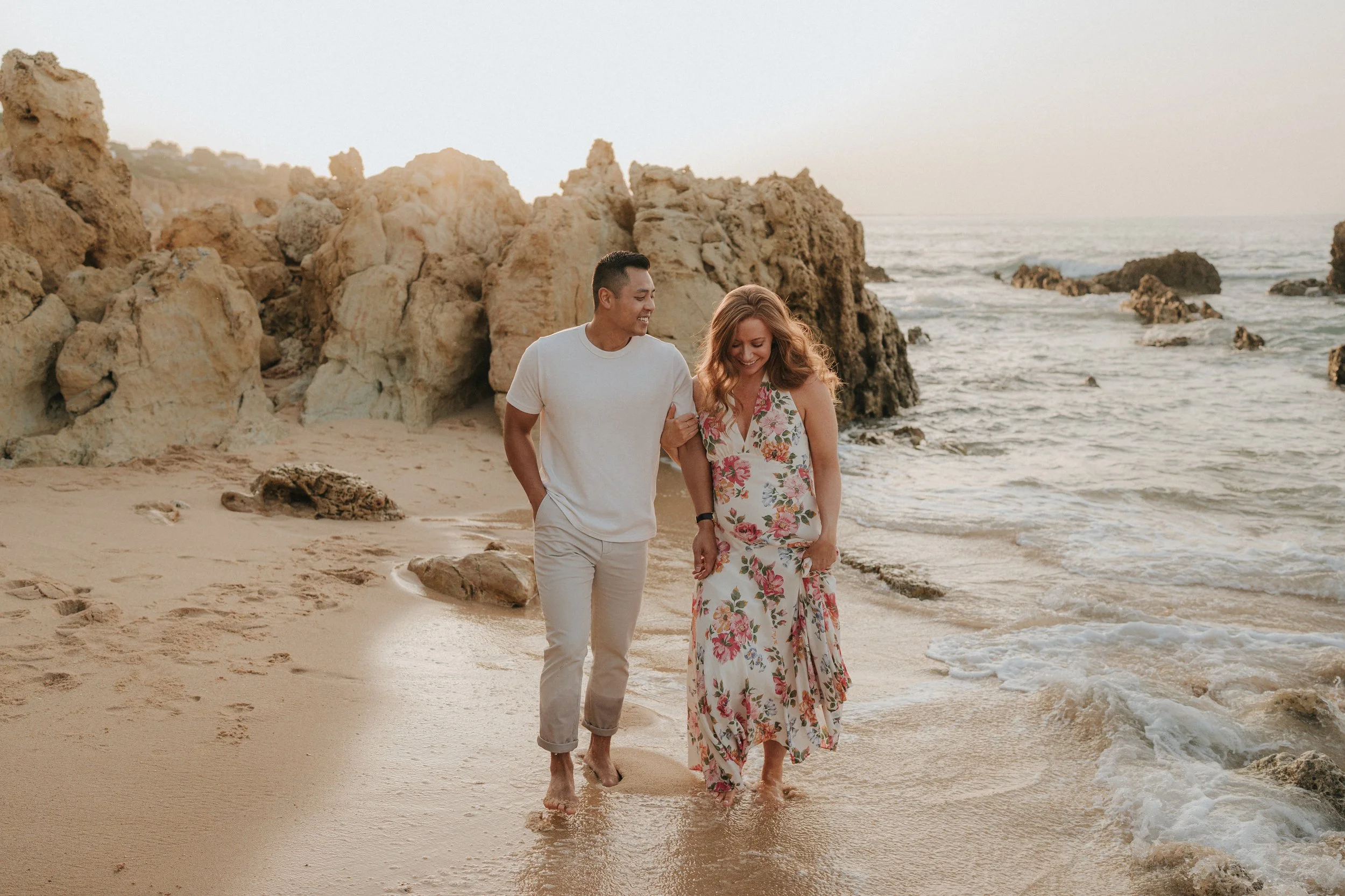 A man and woman walking barefoot on the beach near ocean waves, surrounded by rocks, during sunset. They are smiling and holding hands.