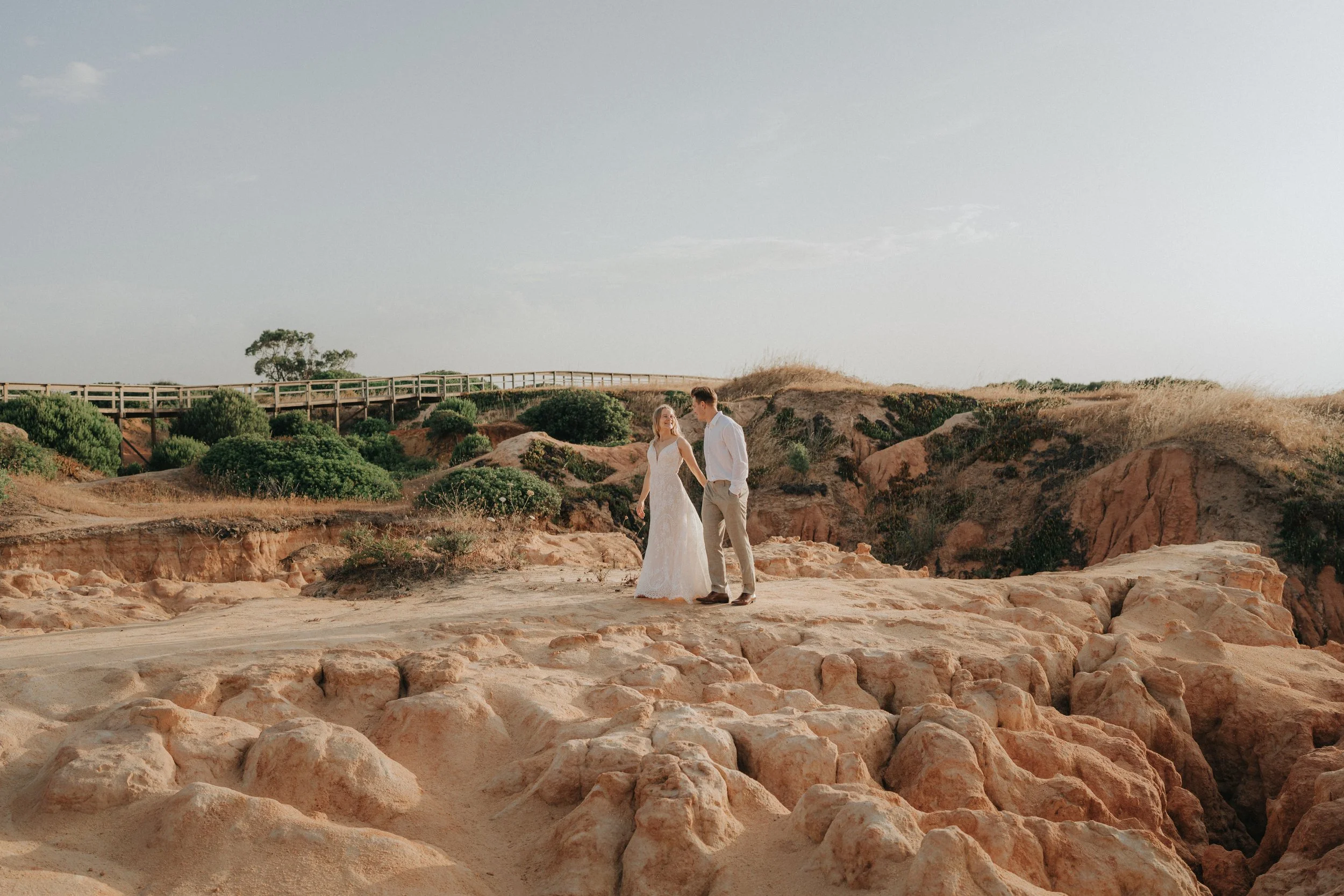 A bride and groom holding hands and looking at each other outdoors on a rocky, sandy terrain with sparse vegetation, a wooden bridge, and a cloudy sky in the background.