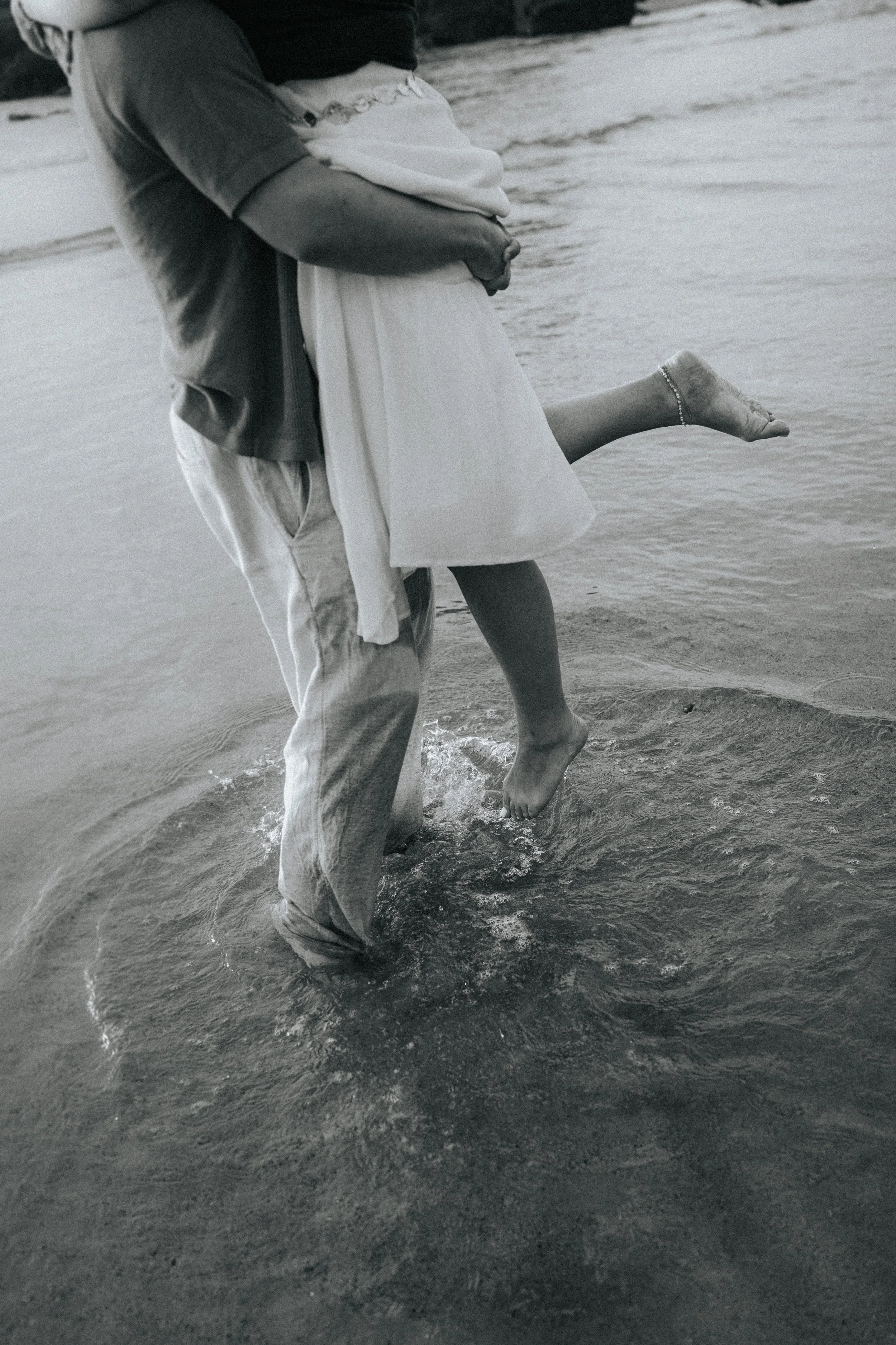 A couple at the beach, with the man holding the woman as she kicks her leg in the air, near the shoreline.