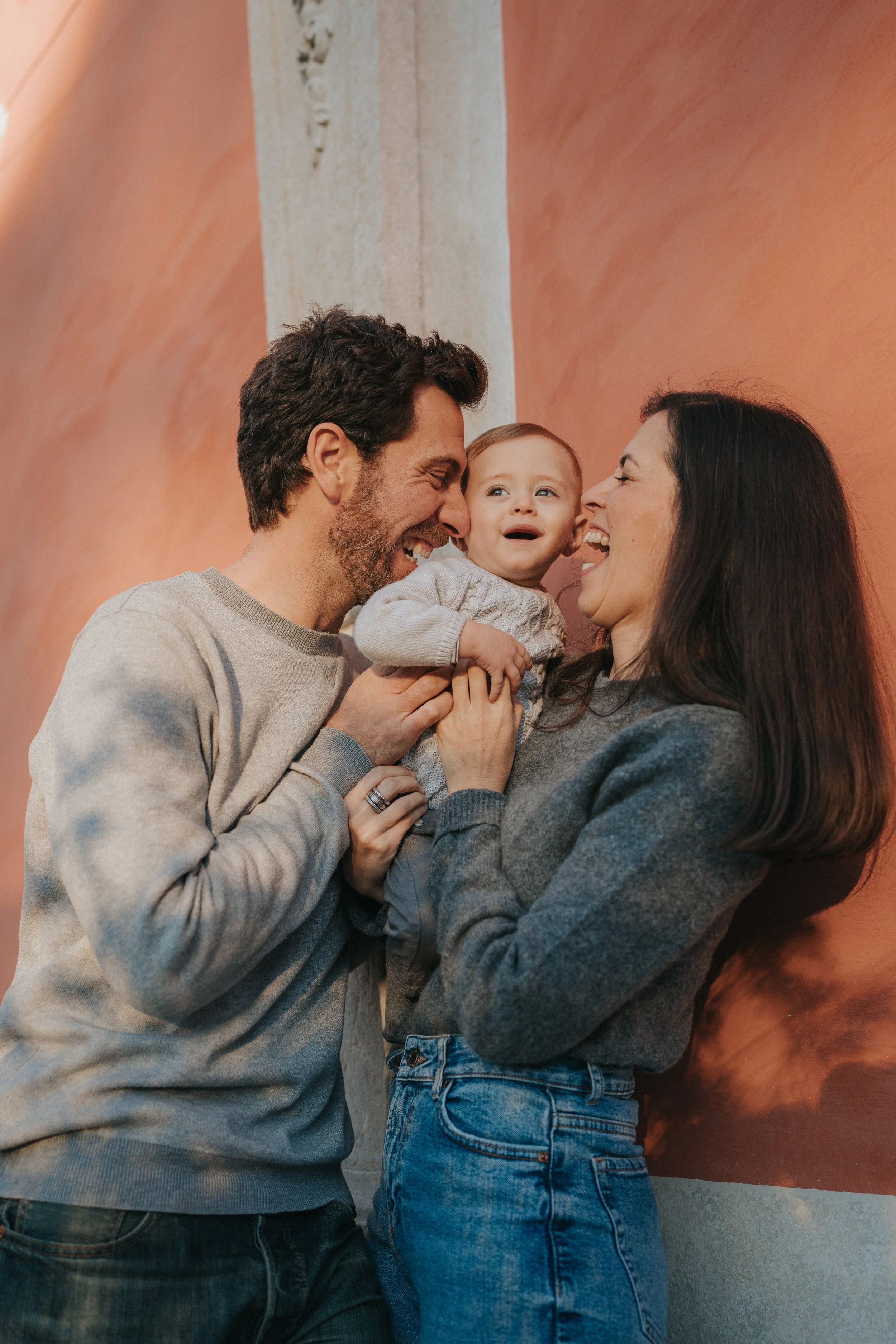 A family of three holding a crying toddler, smiling and laughing together indoors.
