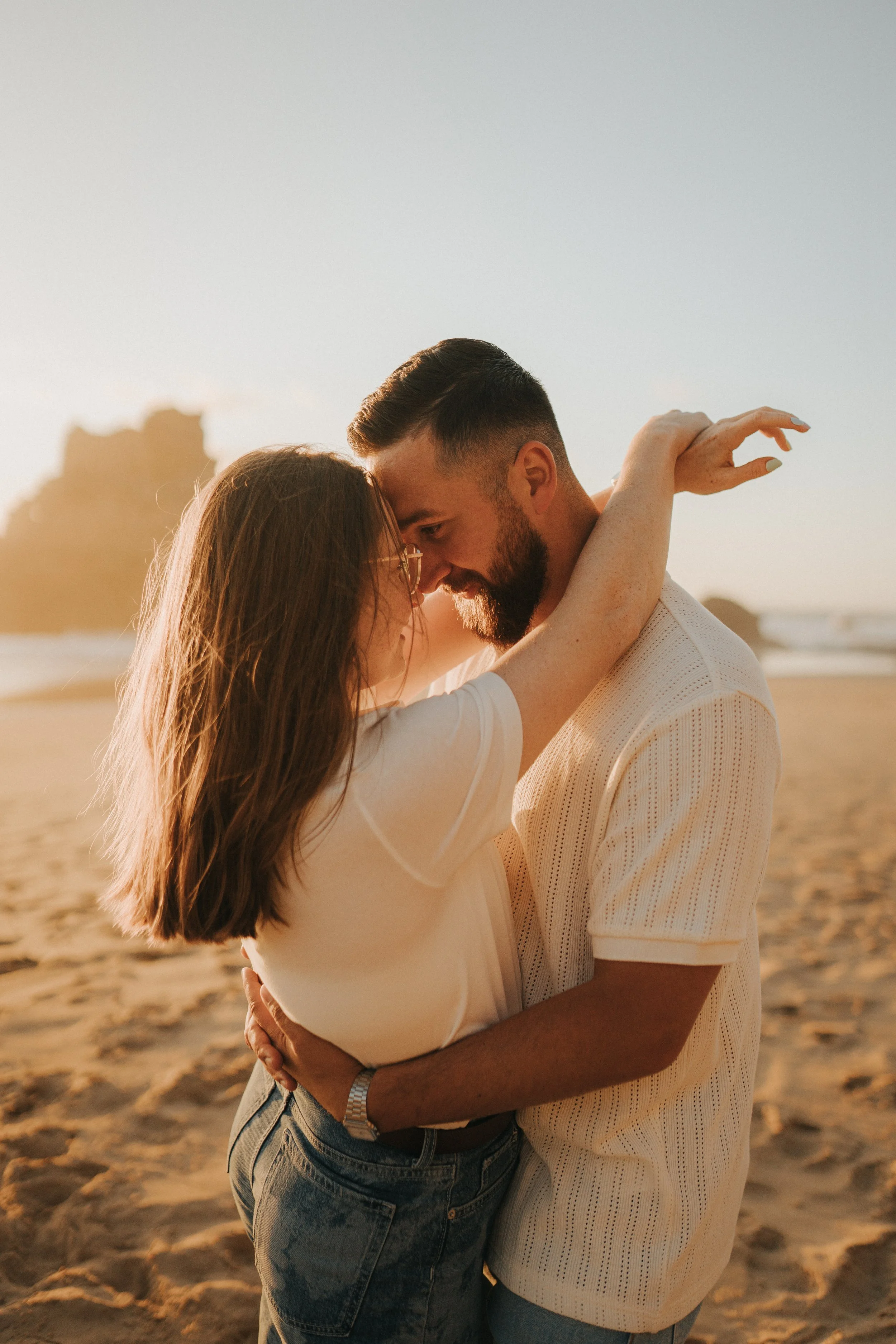 A couple embracing tightly on a beach during sunset, with rocky formations and the ocean in the background.