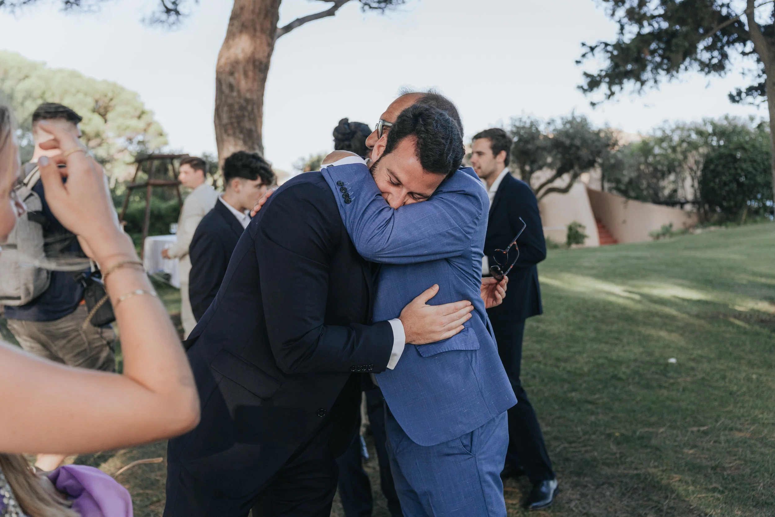 Two men in suits hugging each other outdoors during a gathering, surrounded by other people, under large trees with a grassy area.
