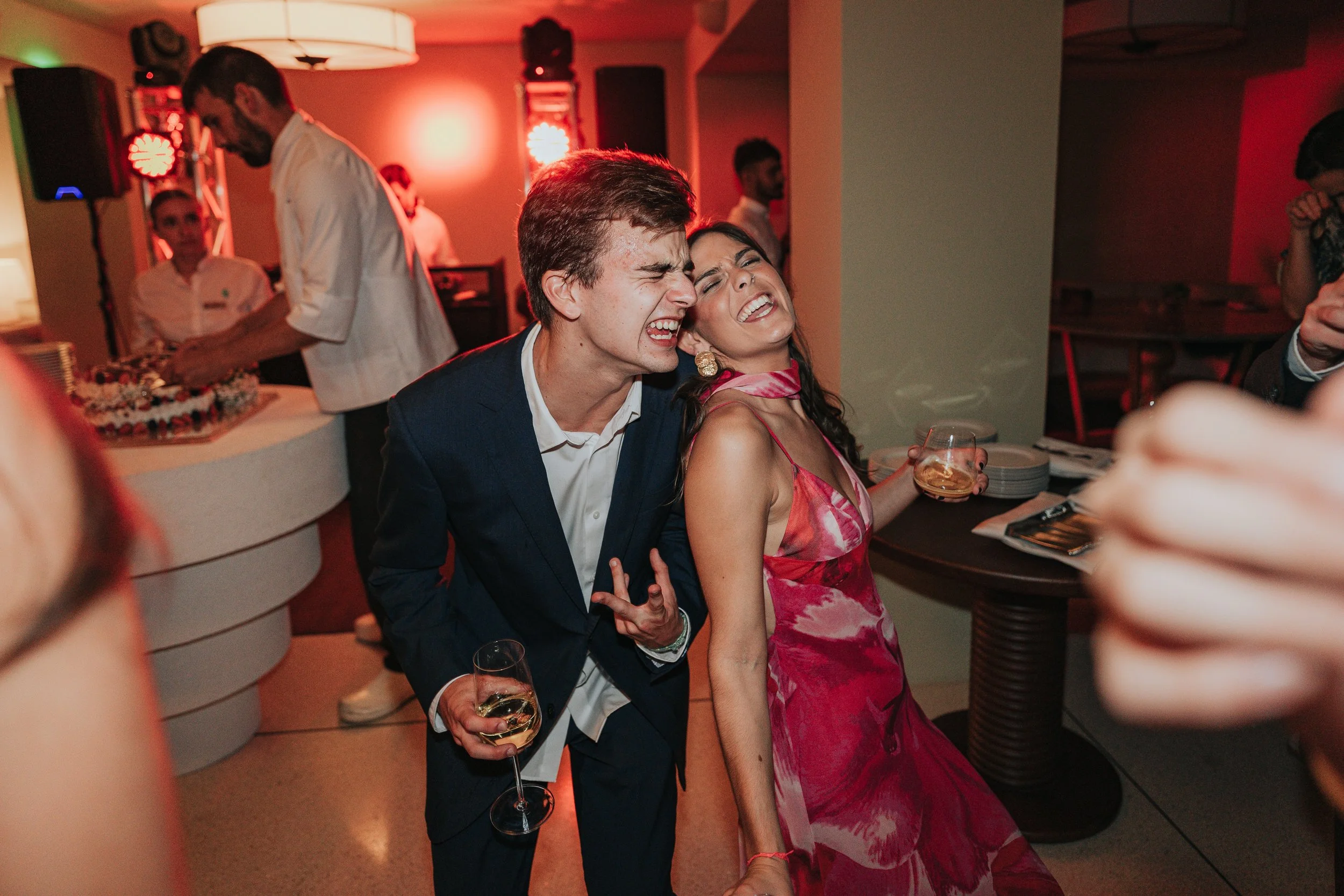 People celebrating at a party, two individuals in the foreground laughing and holding wine glasses, with a cake and DJ setup in the background.