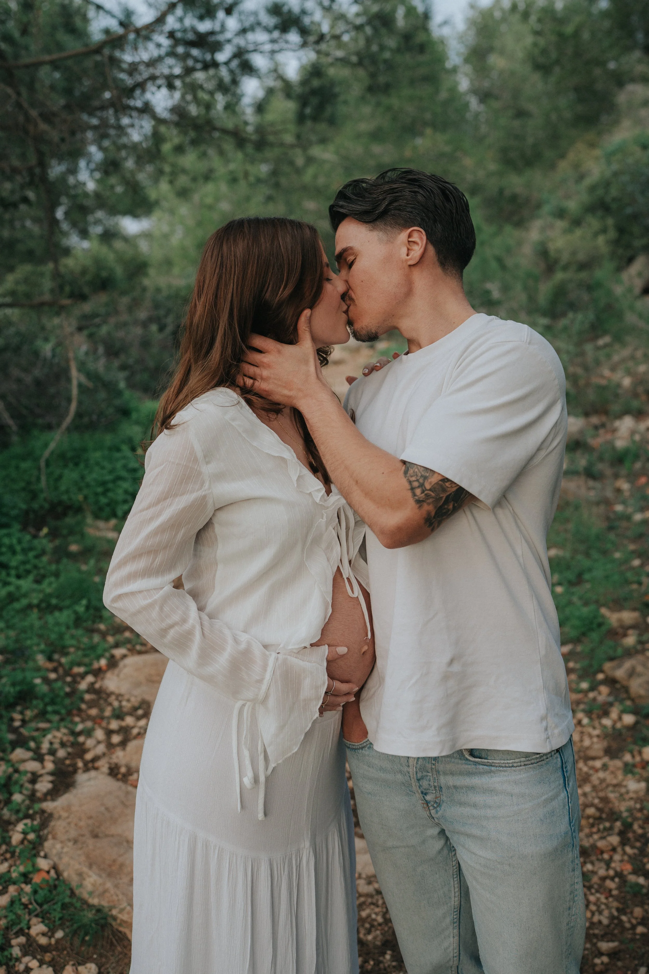 A couple kissing outdoors, with the woman gently touching the man's face and her hand on her pregnant belly. They are surrounded by green trees and plants.