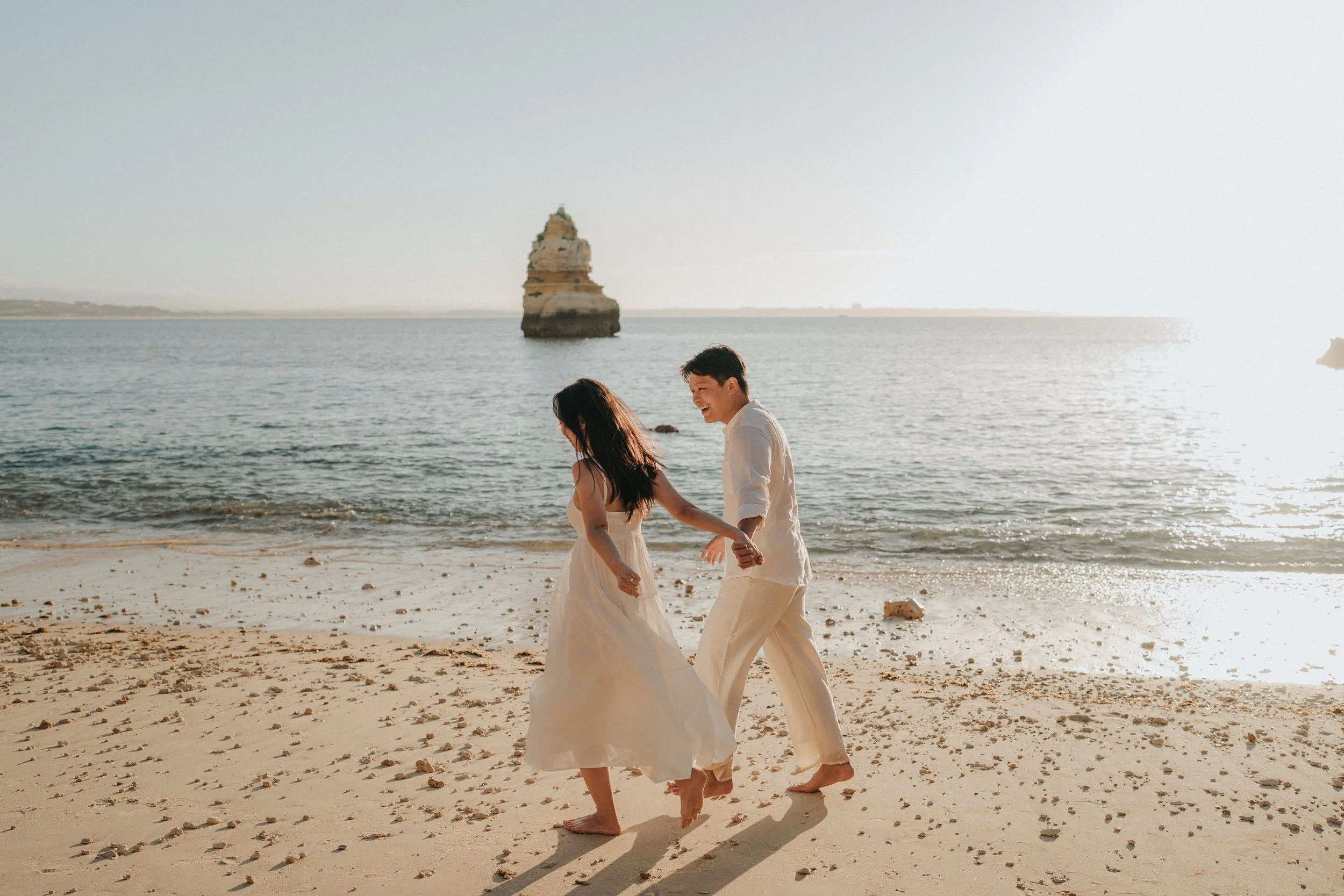 A couple dressed in light-colored clothing holding hands and walking barefoot along the beach during sunset, with a rocky sea stack in the background.