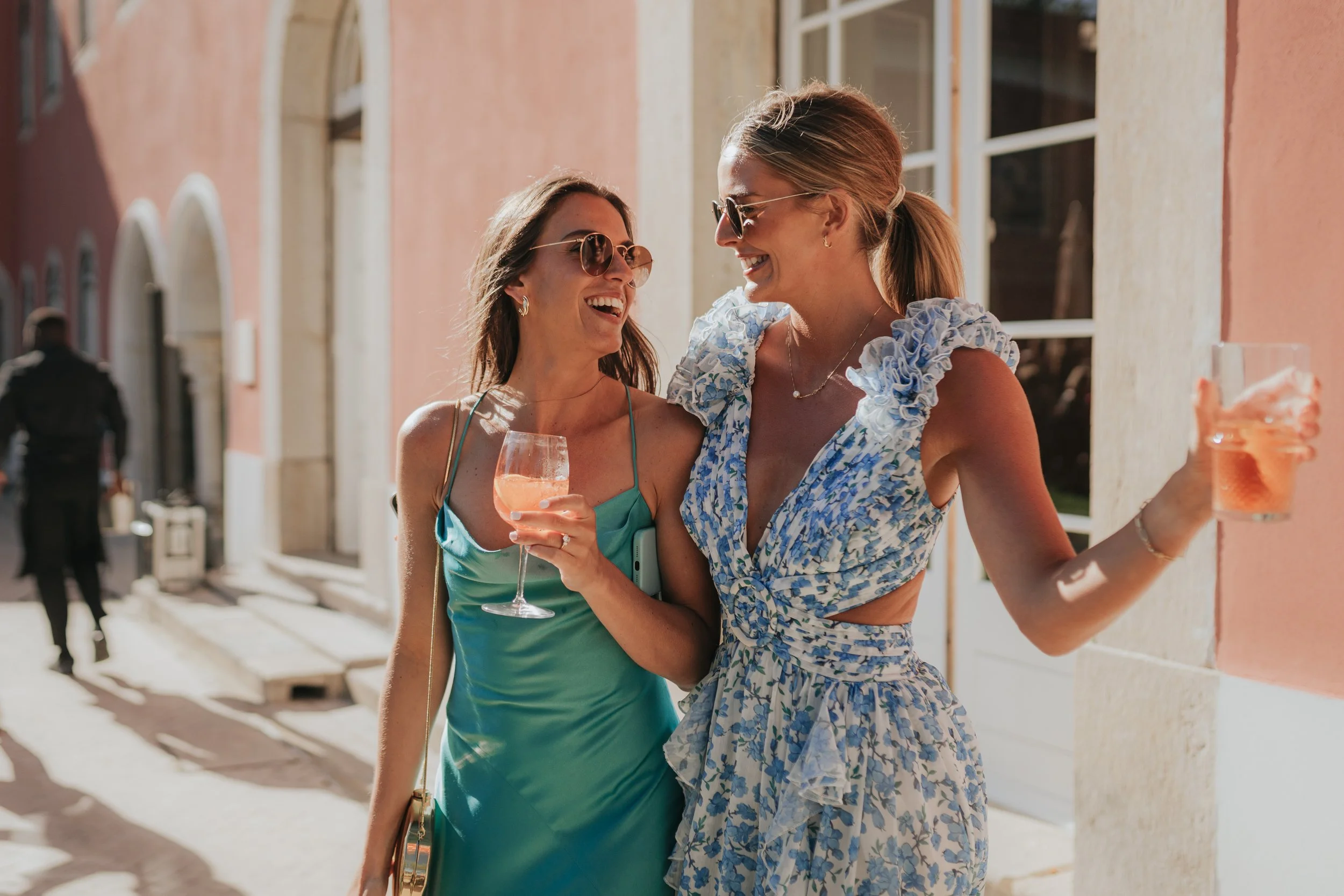 Two women smiling and talking while standing outdoors on a sunny day, one holding a glass of pink drink and the other holding a cup with a pink beverage, both wearing sunglasses and summer dresses.