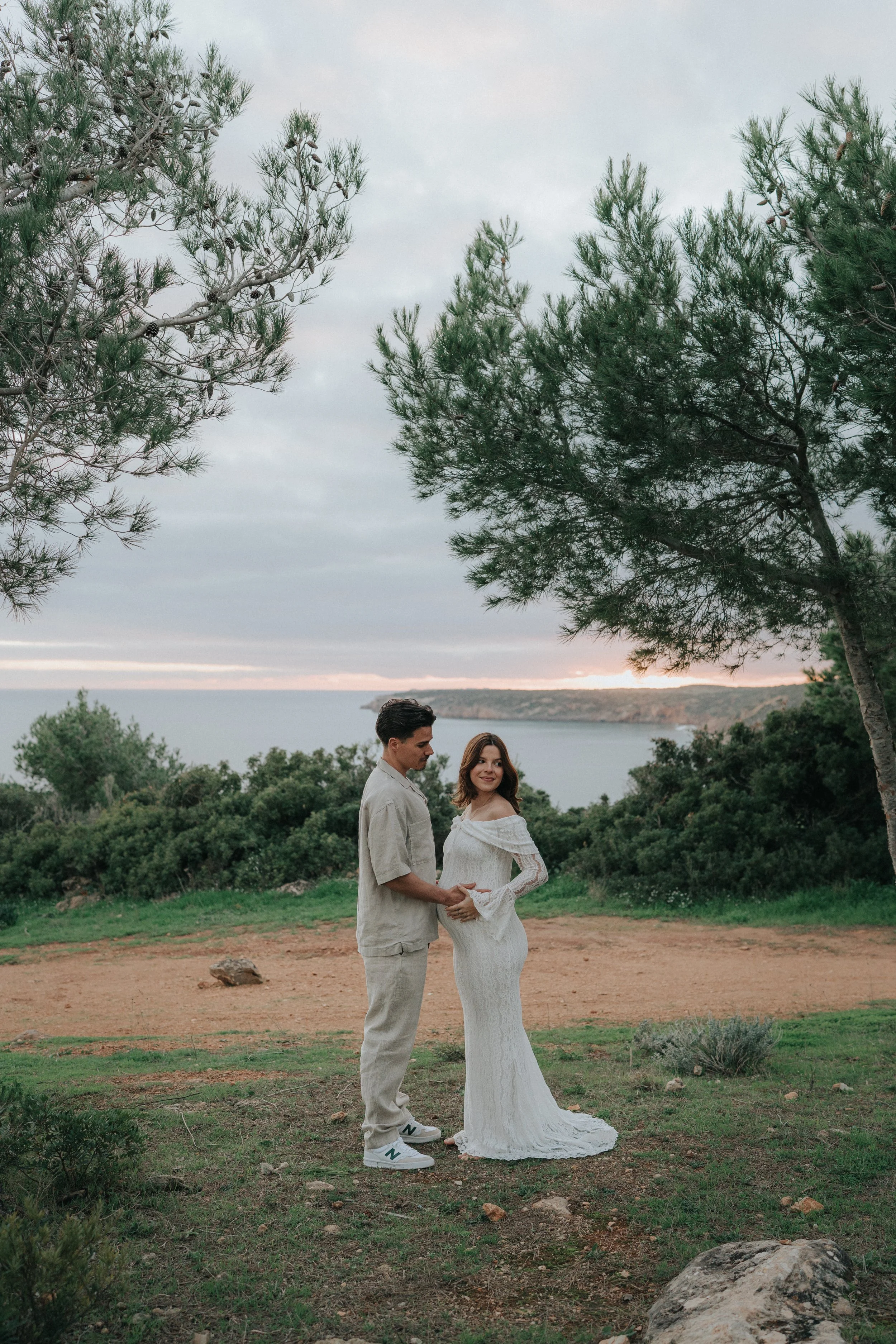 A couple holding hands and looking at each other outdoors during sunset, with trees, bushes, and a body of water in the background.