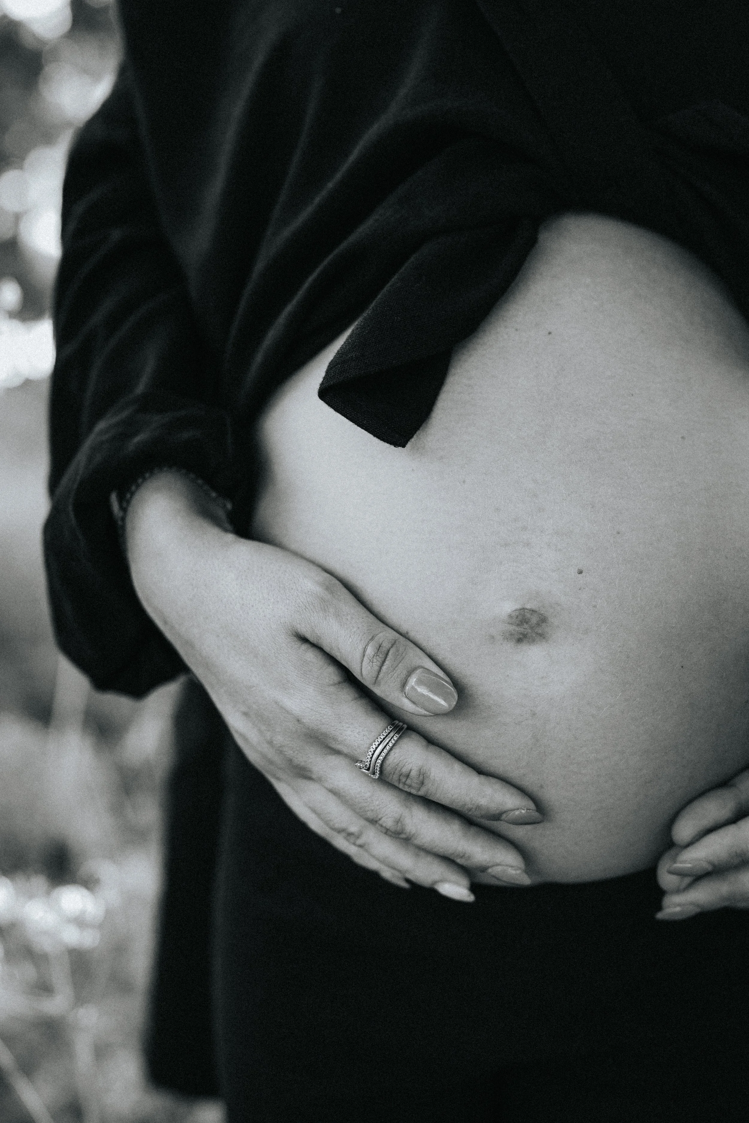 A person, with painted nails and wearing rings, cradles their pregnant belly. They are wearing a dark top with part of their shoulder visible and are outdoors, as indicated by blurred background and light play.