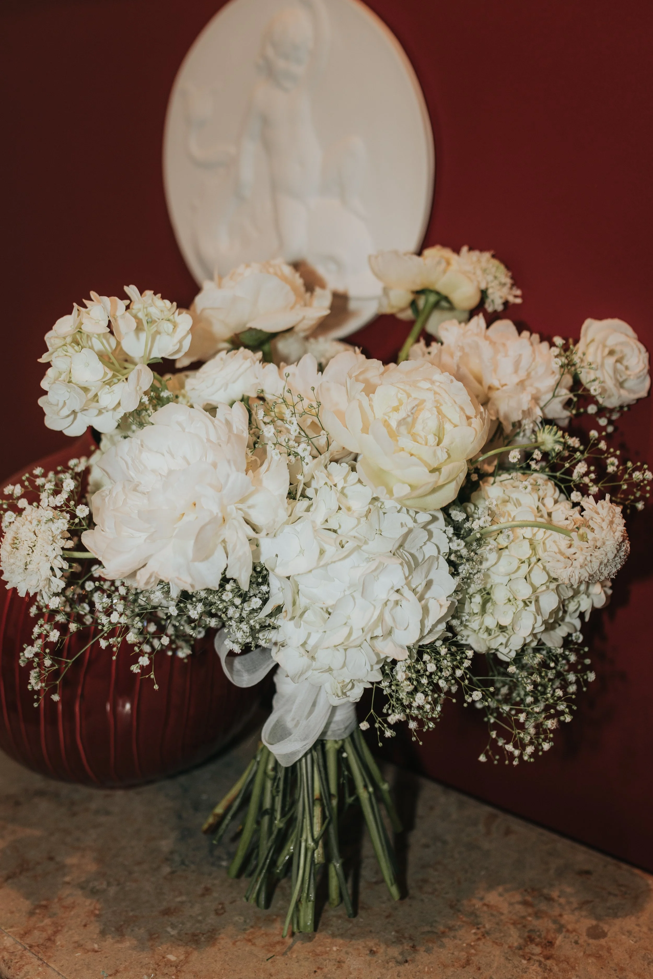 A bouquet of white flowers, including peonies, roses, and baby's breath, resting against a red background with a white medal or plaque featuring a bas-relief of a person on it.
