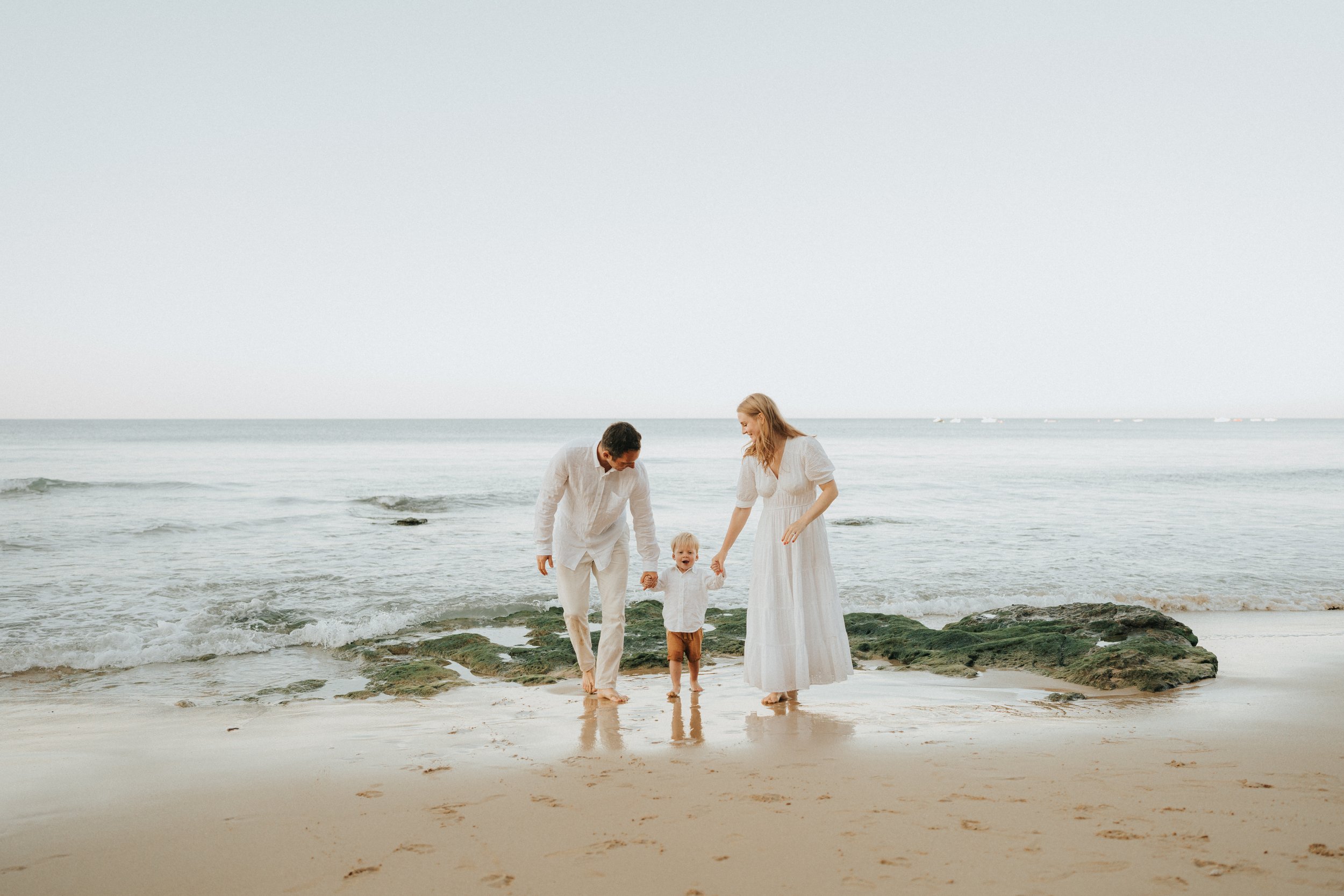 A family of three, a man, woman, and young child, walking hand in hand on a beach with rocks and tide pools during daytime.