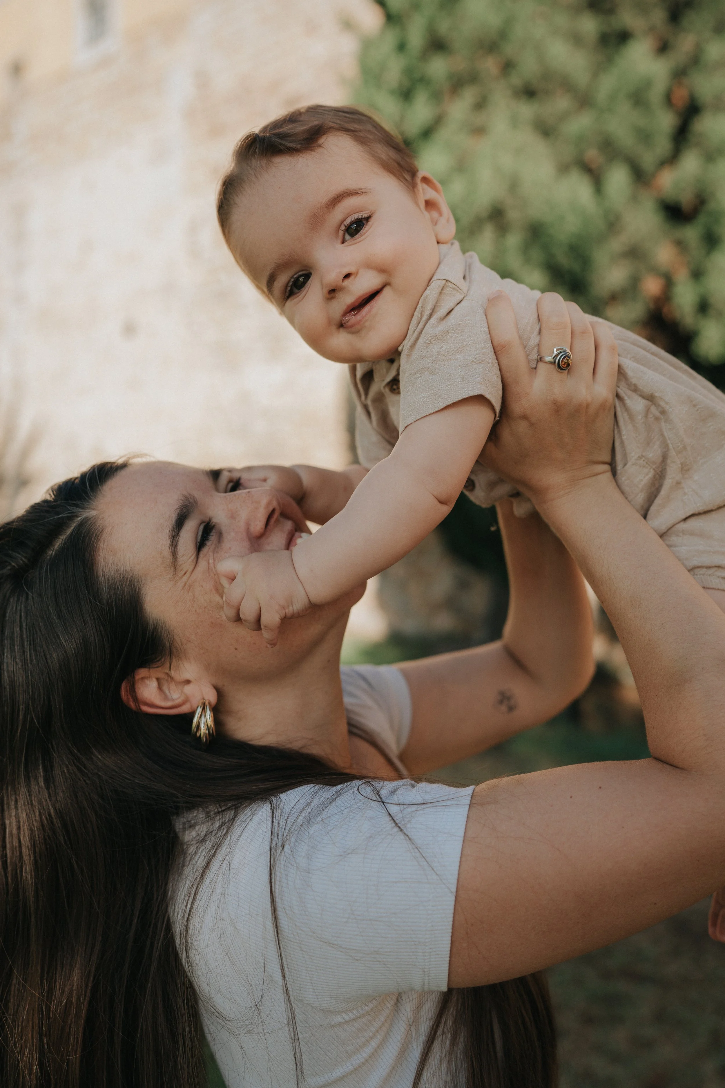 A woman with long dark hair and gold hoop earrings holds a young boy upside down outdoors, both smiling. The boy has short brown hair and is wearing a beige shirt.