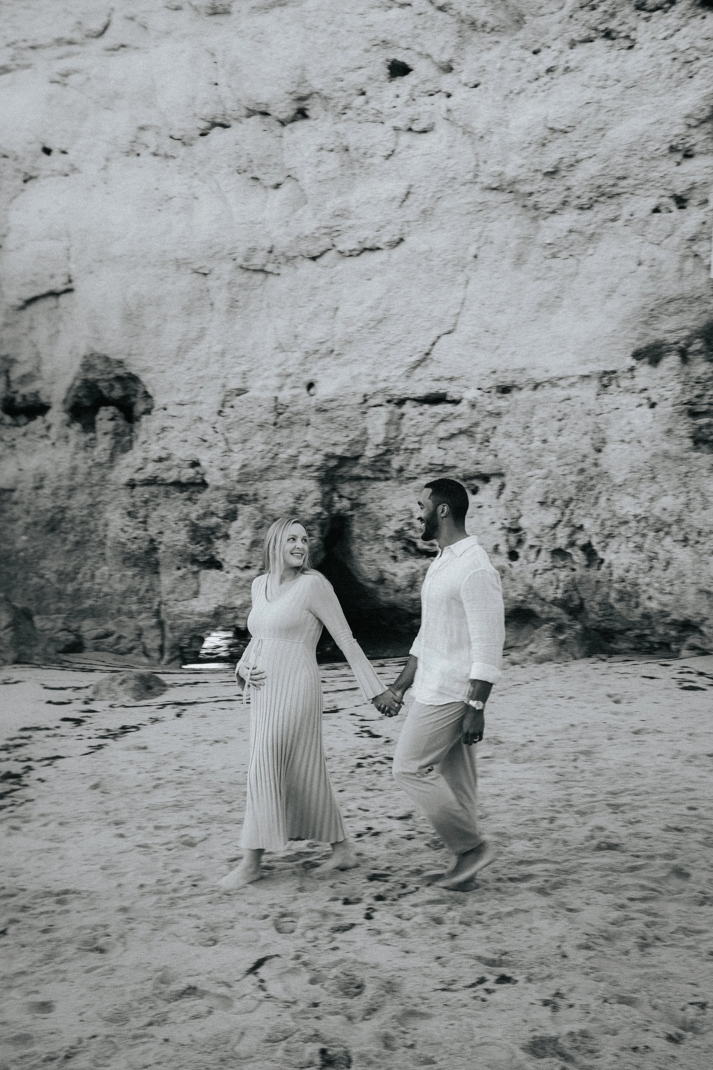 A couple holding hands walking on the beach near rocky cliffs, smiling at each other.
