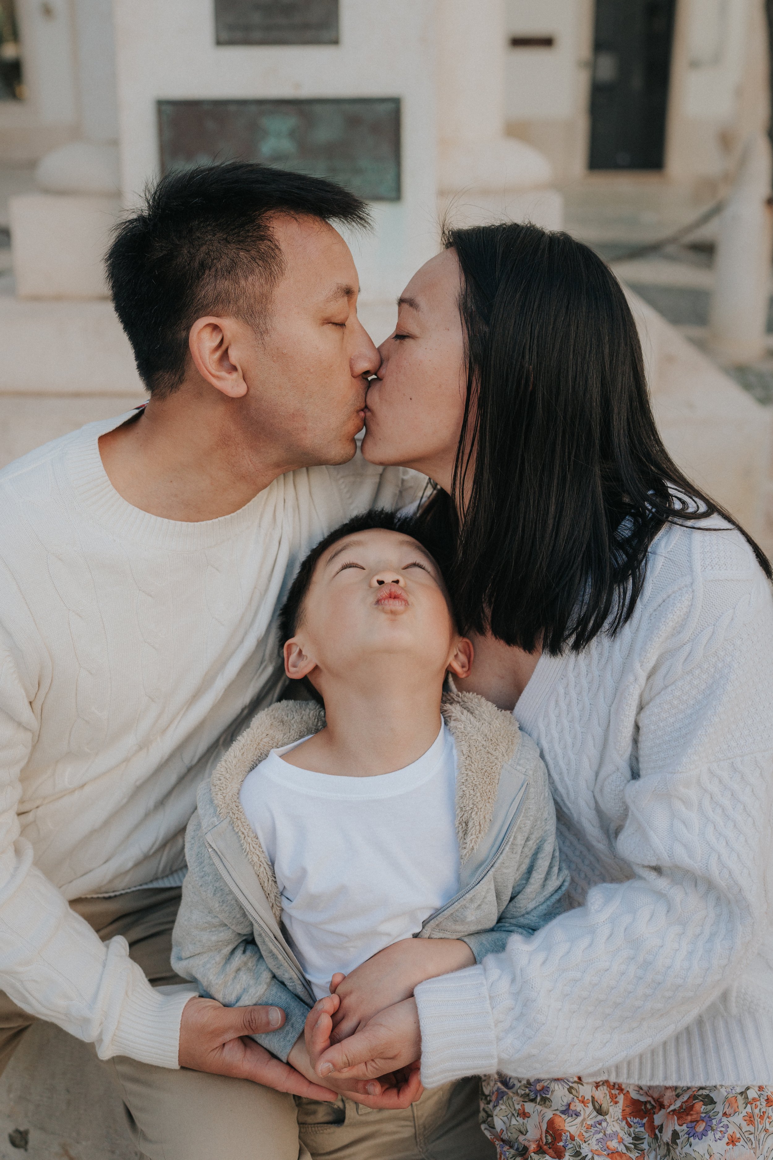 A family of three sharing a kiss, with a young child in the middle, outdoors in front of a building with stairs.