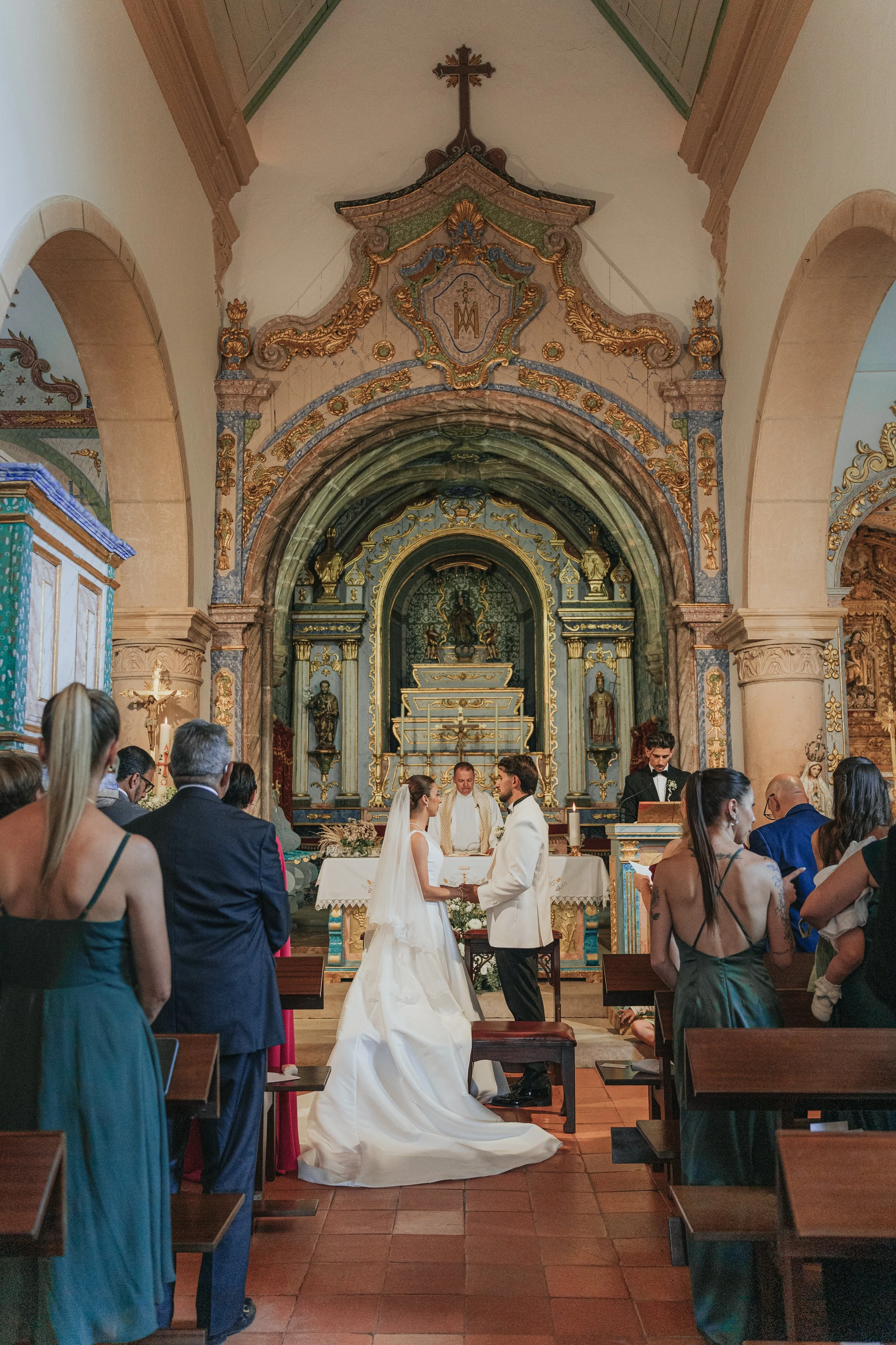 A couple getting married in a church with officiant, surrounded by guests. The interior features ornate decorations, gilded accents, and religious icons.