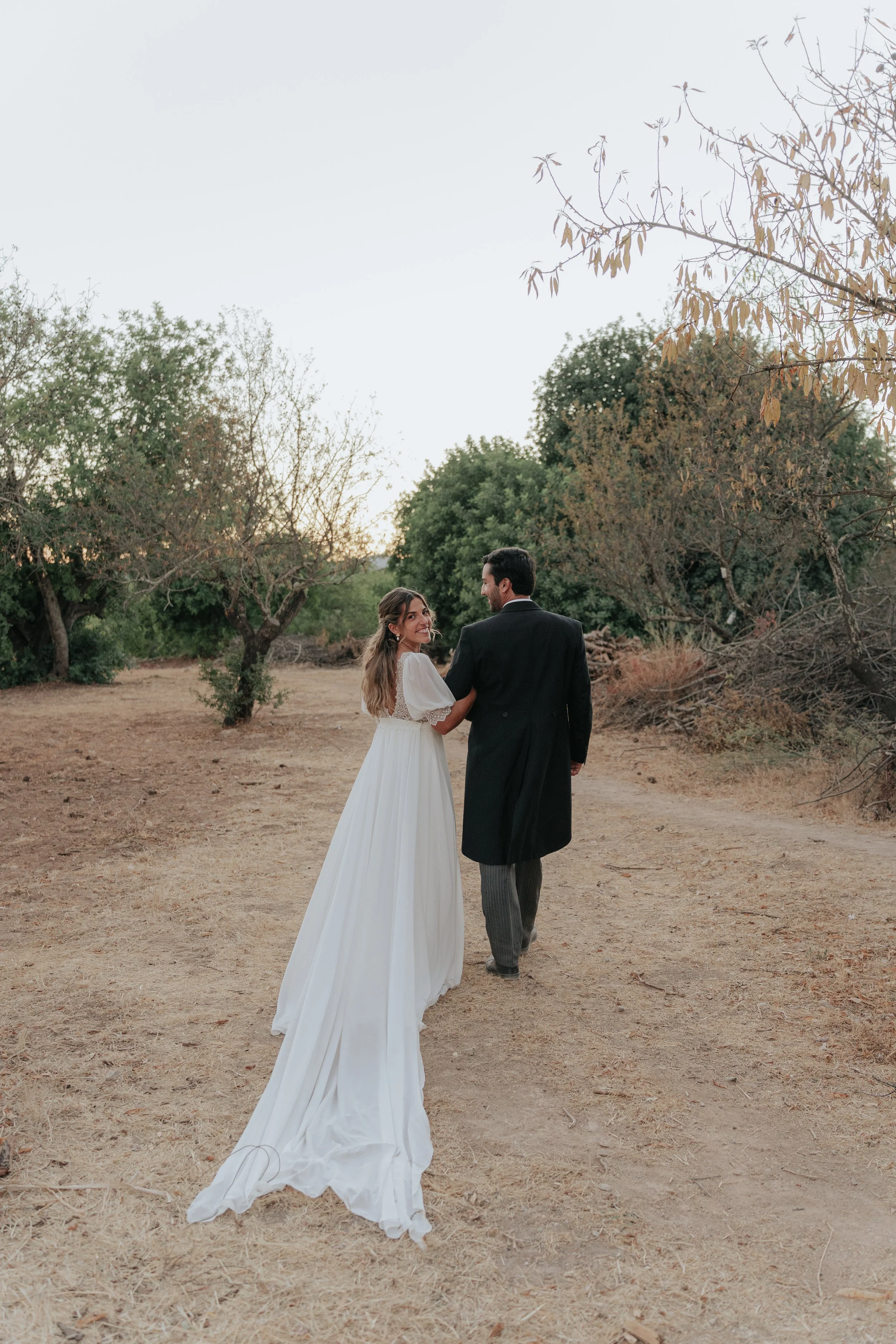 A bride and groom holding hands and walking outdoors in a natural setting with trees and dry grass, as the sun sets in the background.