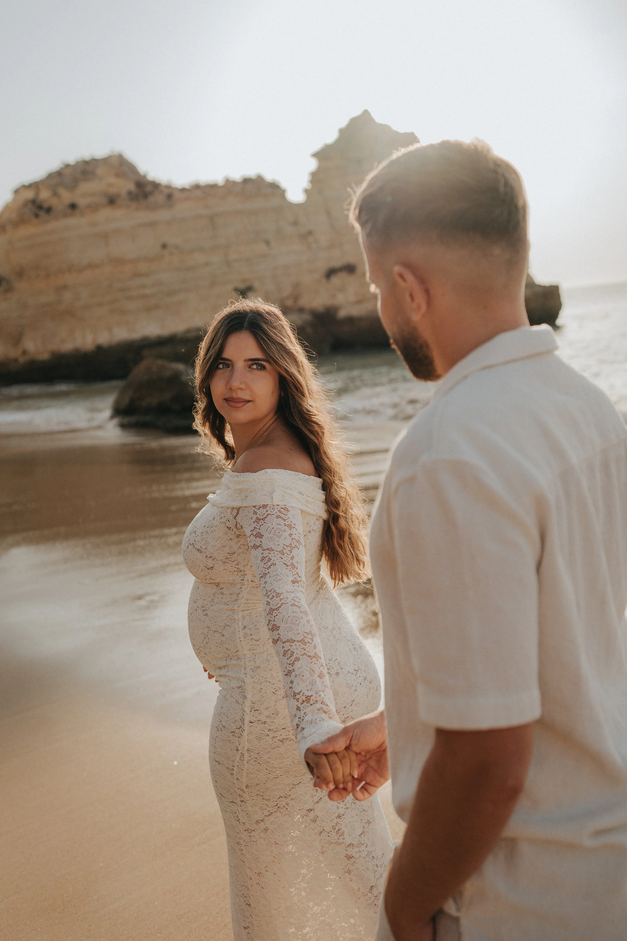 A pregnant woman and a man hold hands on the beach at sunset, with rocky cliffs in the background.