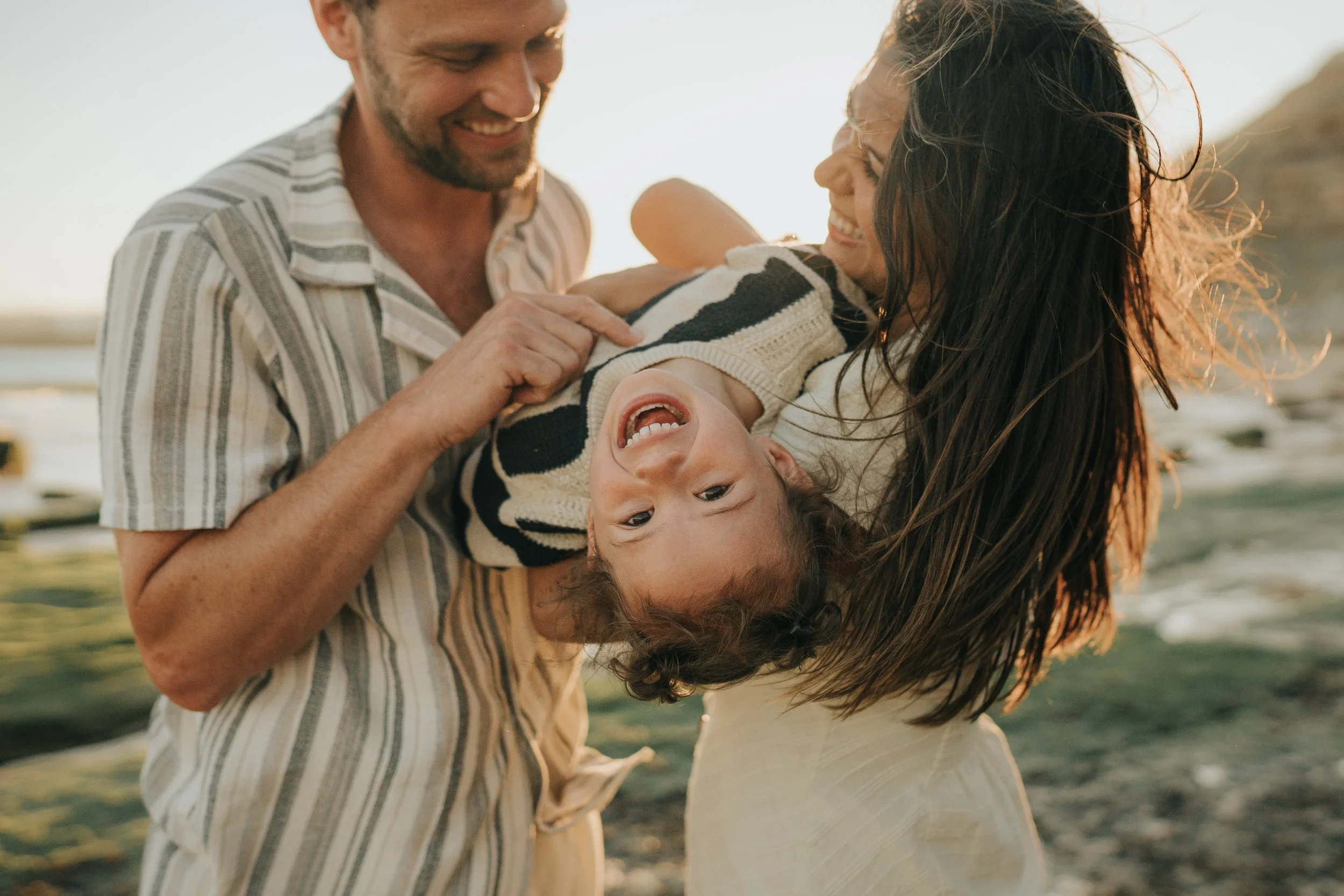 A happy family of three at the beach, with a man, a woman, and a young girl, all smiling and enjoying each other's company during sunset.
