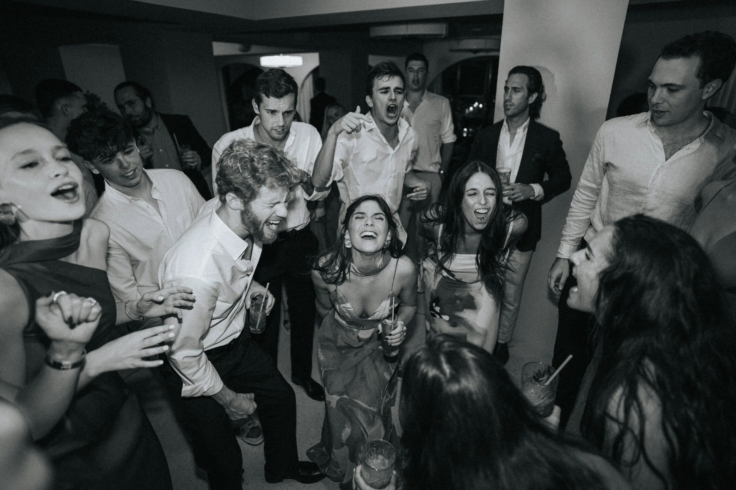 A group of people at a party or celebration, mostly young adults, are laughing, singing, and enjoying drinks in a social setting.