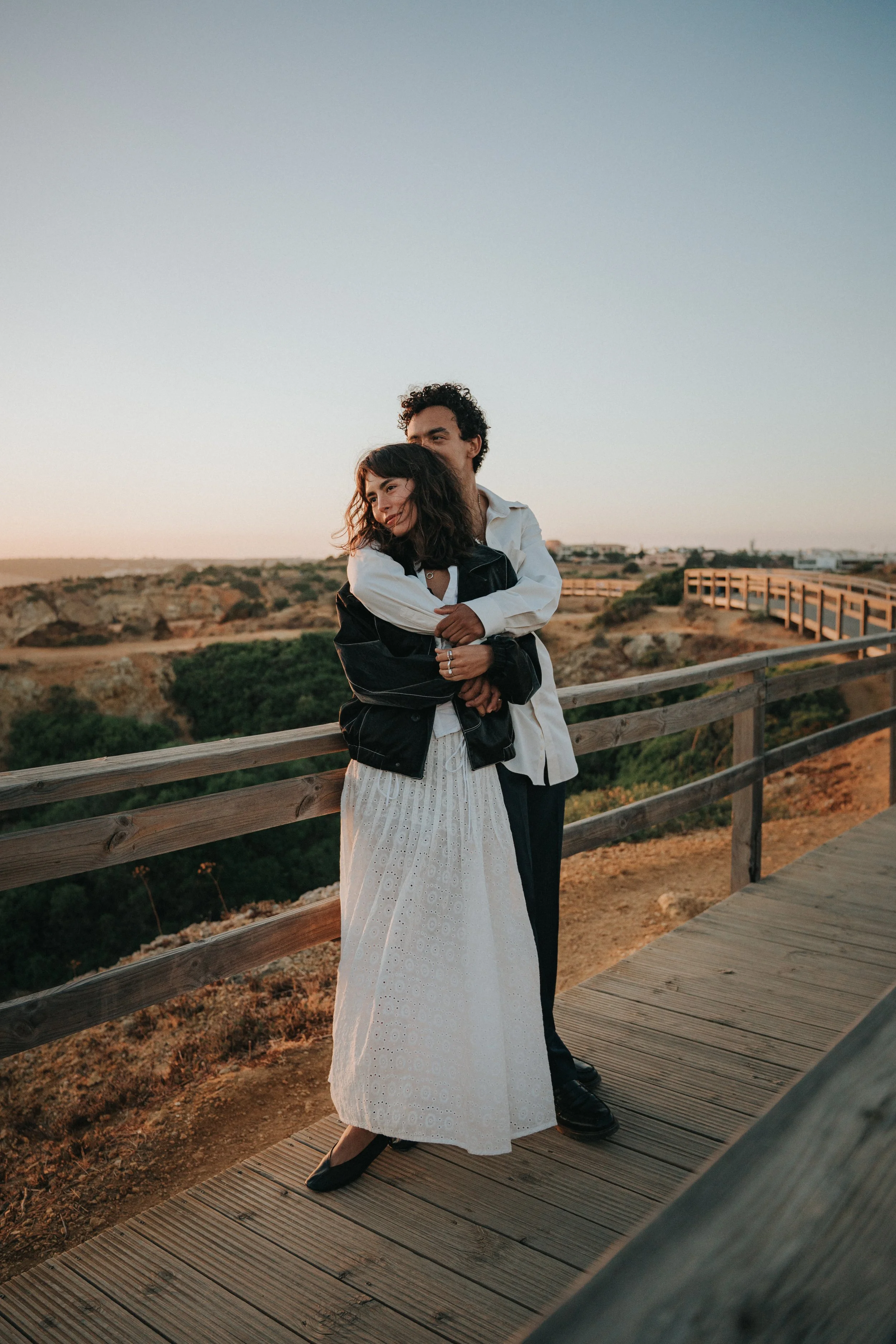 A couple standing on a wooden walkway outdoors during sunset, the woman is leaning into the man, smiling softly, and he is standing behind her with his arms around her shoulders, both appearing happy and close.