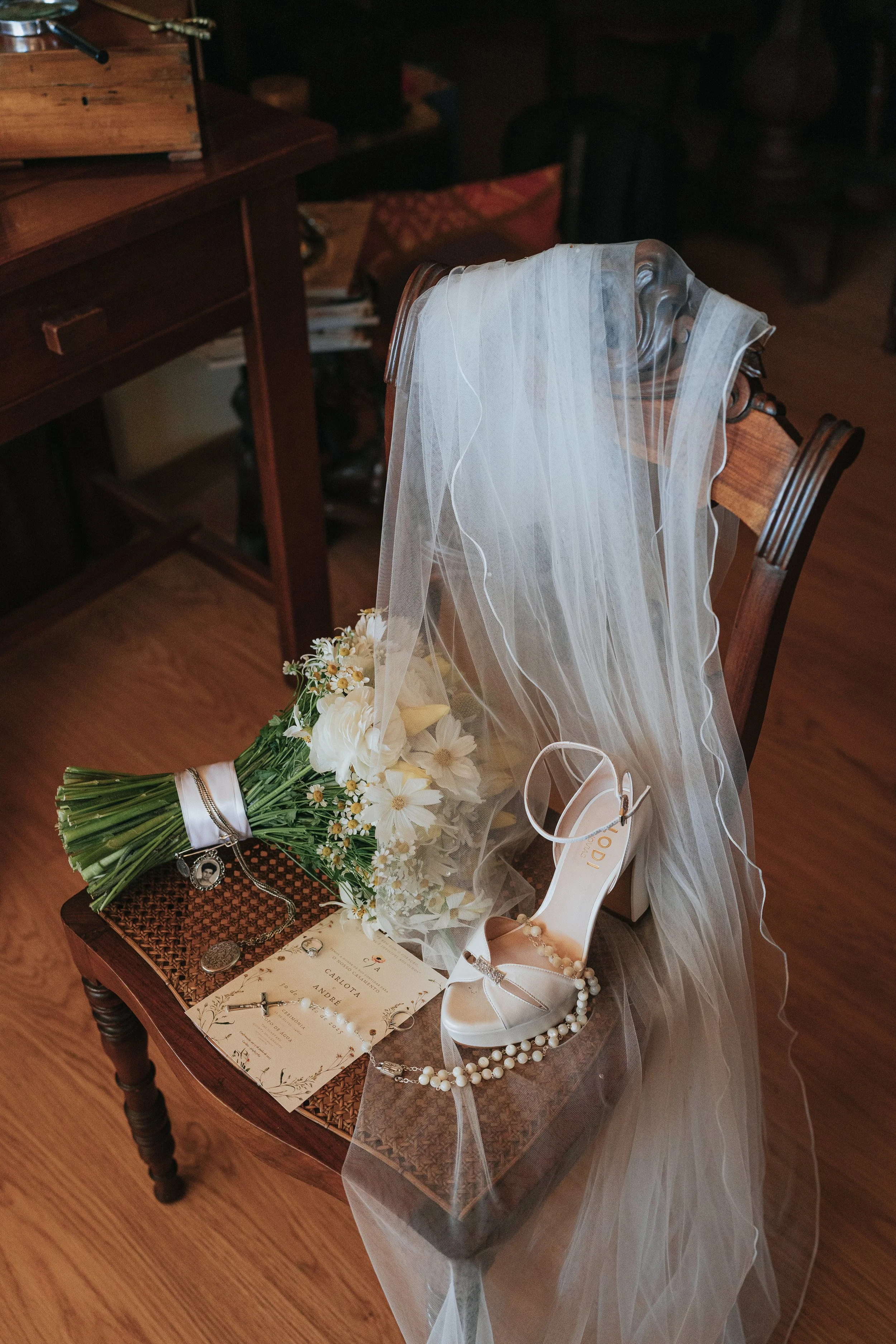 A table with a wedding veil, a bouquet of white flowers, a pair of white high-heeled shoes, a wedding invite, jewelry, and a small photo frame.