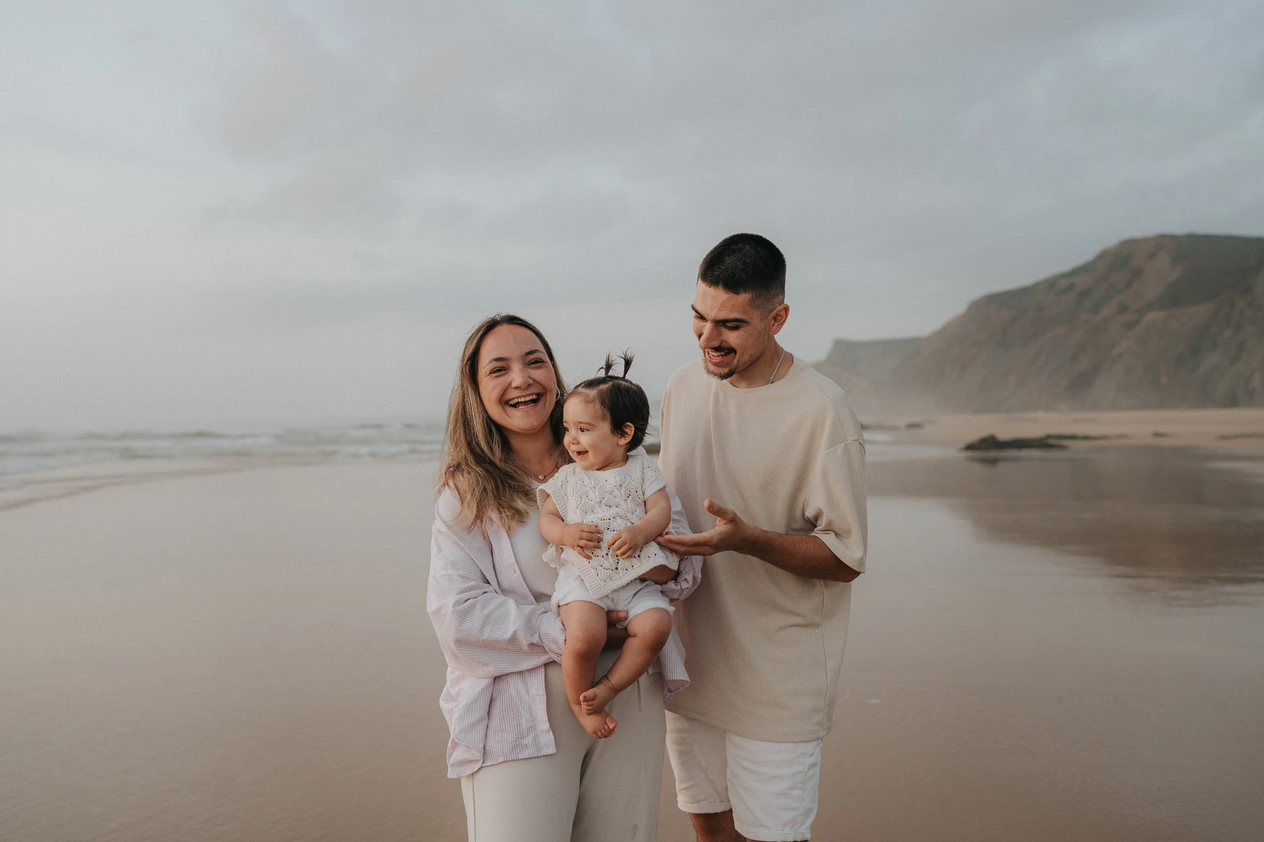 A happy family of three at the beach, with a woman holding a young child and a man standing beside them, all smiling and enjoying a cloudy day.