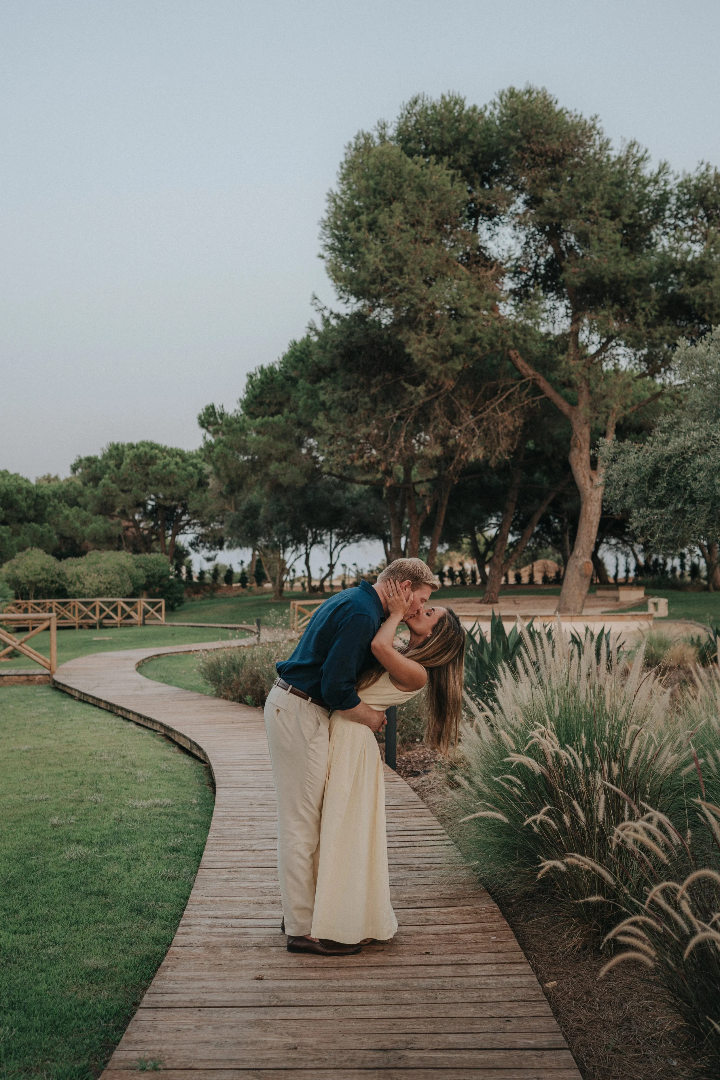 A couple sharing a kiss on a wooden walkway in a park with lush greenery and tall trees in the background, during the daytime.