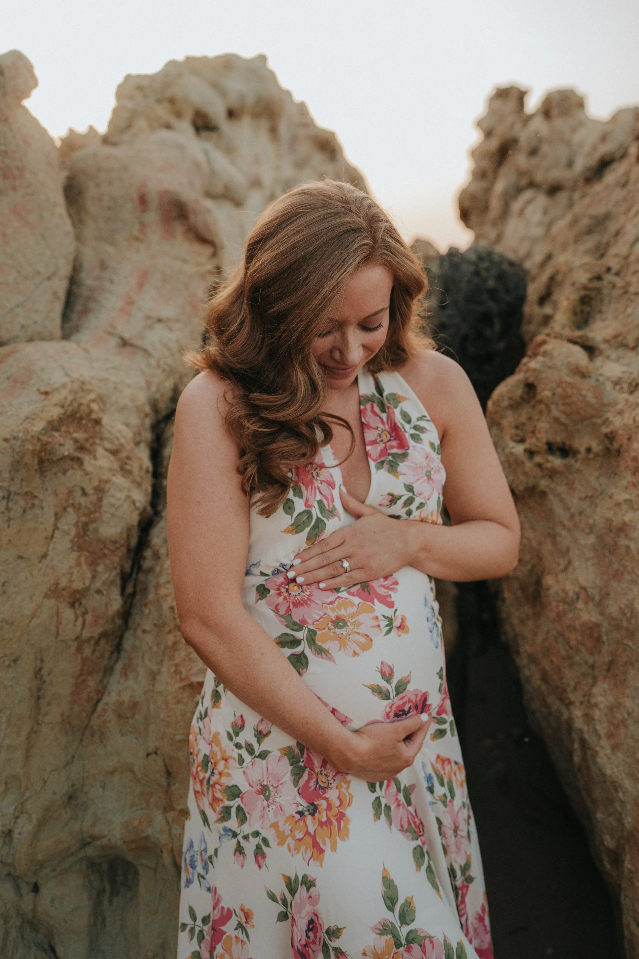 A woman in a floral dress holding her pregnant belly, standing amidst large rock formations.